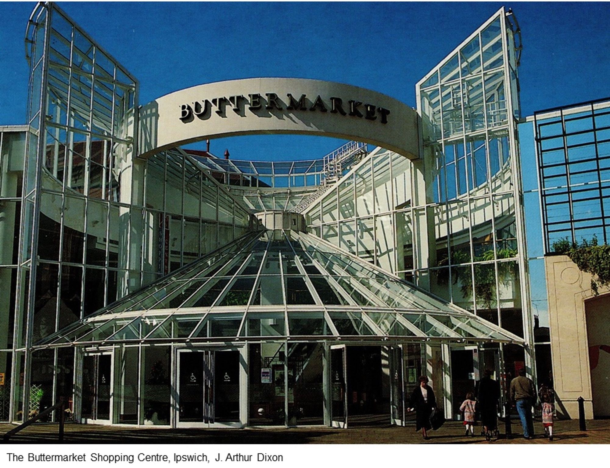 The Buttermarket Shopping Centre, Ipswich. The entrance sits under a glass conical roof. Either side of the entrance are decorative “wings” in steel and glass. A J. Arthur Dixon postcard