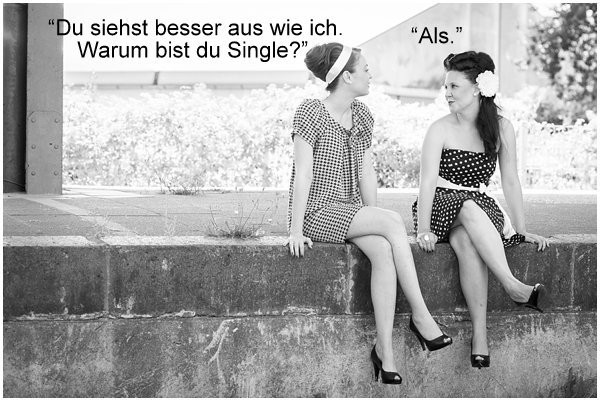 Auf dem schwarz-weißen Foto sitzen zwei Frauen nebeneinander auf einer niedrigen Mauer oder Kante im Freien.

* Beide tragen kurze Kleider mit Muster (gepunktet bzw. kariert) und hohe Schuhe.
* Die Frau links hat ein Haarband im Haar, die Frau rechts eine große Blume als Haarschmuck.
* Sie schauen sich gegenseitig an, als wären sie im Gespräch.

Oben im Bild ist ein Text eingefügt:
Links steht: **„Du siehst besser aus wie ich. Warum bist du Single?“**
Rechts steht als Antwort: „Als.“

