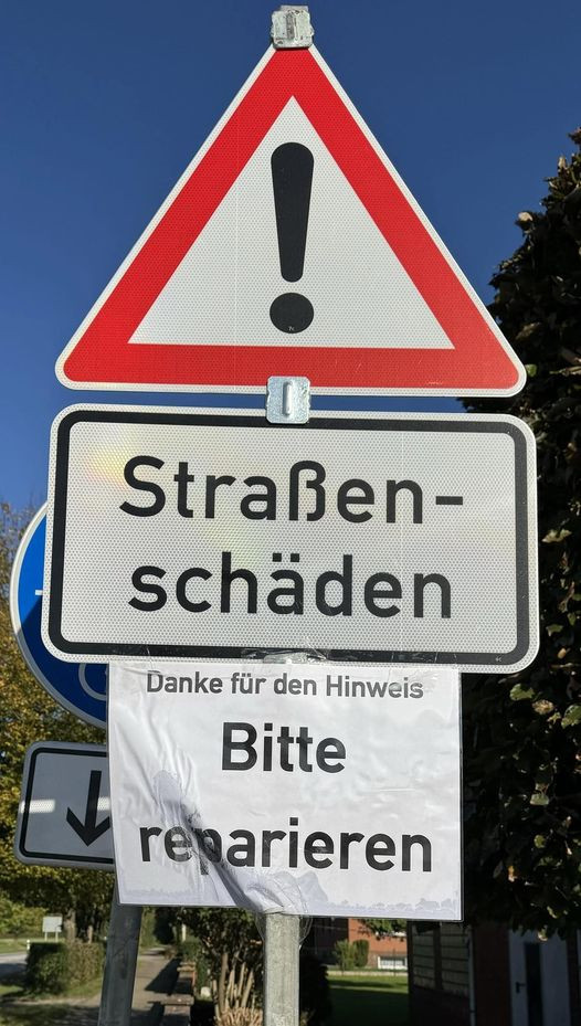 Das Bild zeigt ein Verkehrsschild mit einem roten Dreieck und einem schwarzen Ausrufezeichen in der Mitte, das auf eine Gefahr hinweist. Darunter befindet sich ein weiteres Schild mit der Aufschrift "Straßenschäden", was auf Schäden an der Fahrbahn hinweist. An diesem Schild ist zusätzlich ein Papier befestigt, auf dem steht: "Danke für den Hinweis. Bitte reparieren." 