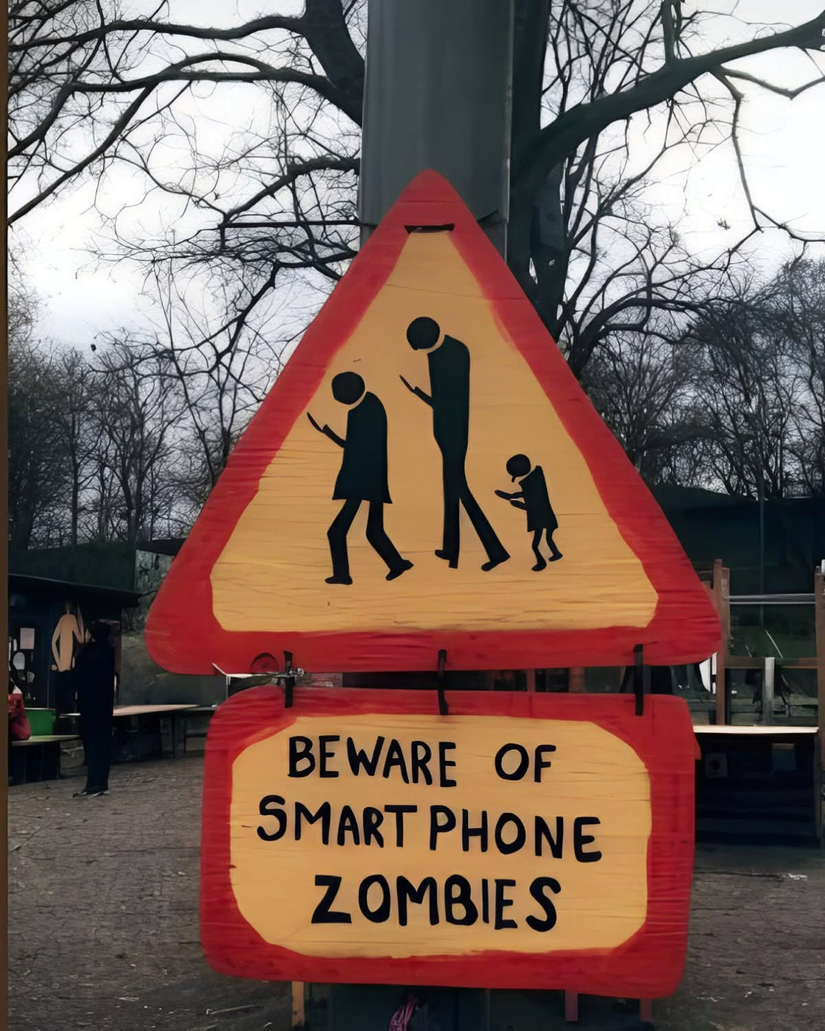 Auf dem Bild ist ein selbstgemaltes Schild zu sehen.

Das obere Schild ist dreieckig, gelb mit rotem Rand, ähnlich einem Verkehrsschild. Darauf sind drei schwarze Silhouetten von Personen dargestellt – ein Erwachsener, ein Jugendlicher und ein Kind. Alle Figuren gehen nach vorne gebeugt und schauen auf ein Smartphone in ihren Händen.

Darunter hängt ein rechteckiges Schild, ebenfalls gelb mit rotem Rand. Darauf steht in schwarzen, handgemalten Buchstaben:
"BEWARE OF SMARTPHONE ZOMBIES".

Im Hintergrund sind kahle Bäume, ein grauer Himmel und Teile eines Spielplatzes oder einer Holzkonstruktion zu erkennen.

