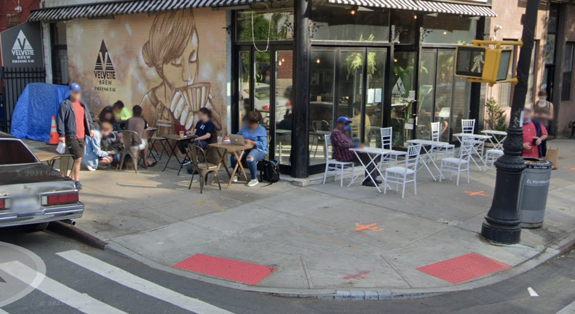 Sidewalk cafe with white tables on one side and more tables on the left with people sitting at them. The wall features a painting of a woman sipping a cup of coffee.