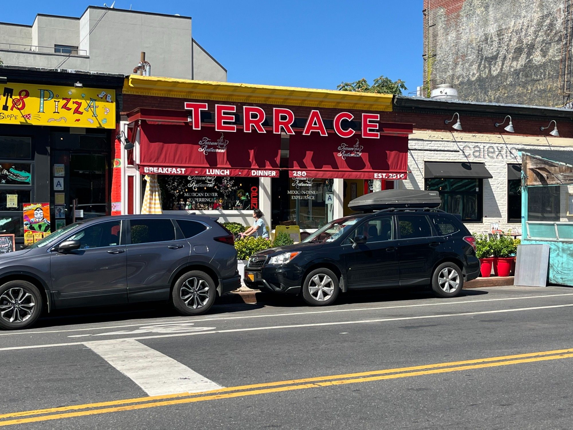 Same city street with three businesses in a one-story building. Two cars are parked in front of a business that has a red sign that reads 'Terrace.'