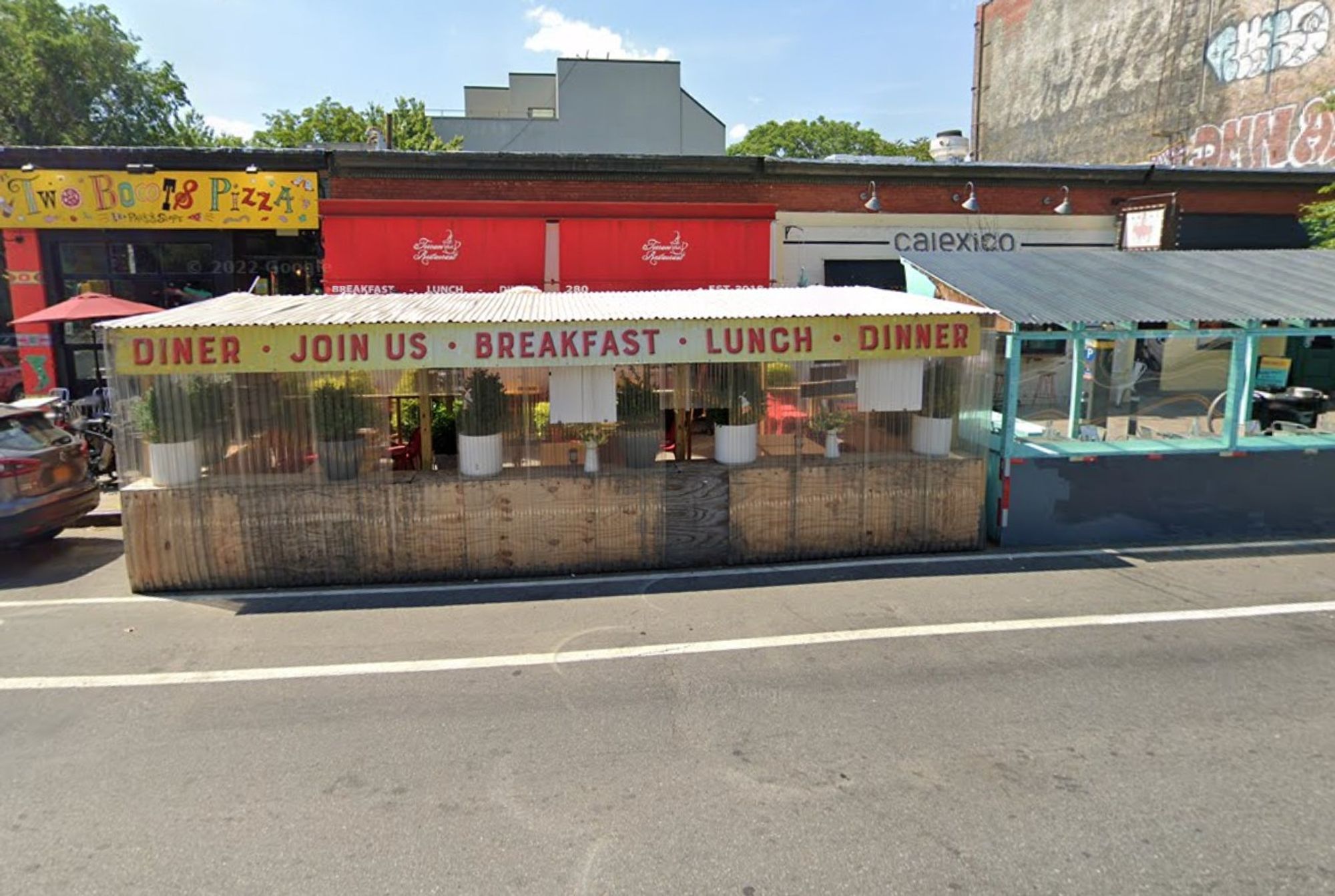 Outdoor dining structure on city street in front of three businesses in a one-story building. Structure has a yellow banner that reads "Diner - Join Us - Breakfast - Lunch - Dinner.