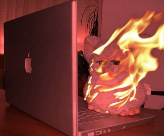 Picture of a Furby toy on yop of a laptop. The picture has a reddish ting, and the Furby is literally on fire.