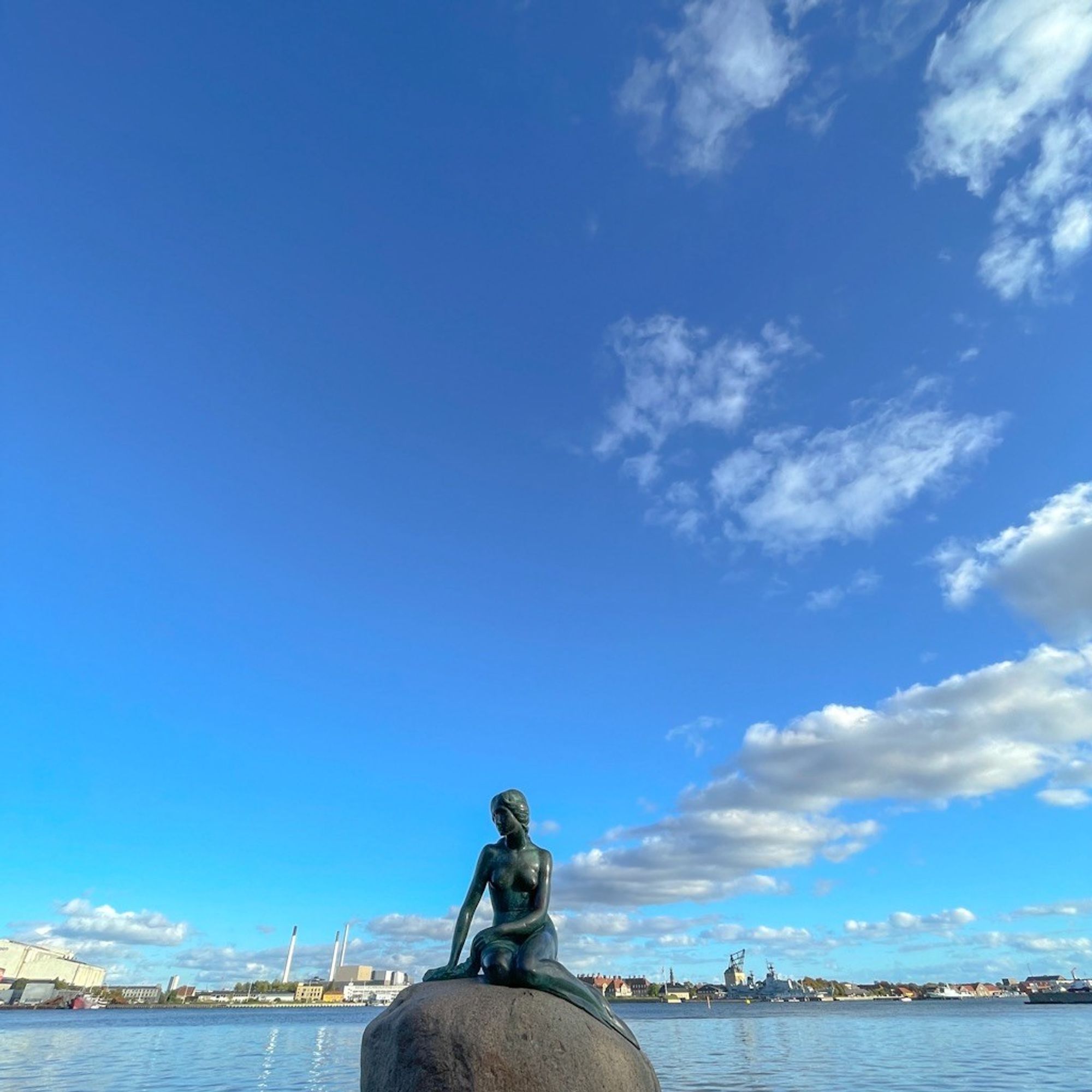 Eine Bronzestatue der kleinen Meerjungfrau auf ihrem Felsen am Wasser, überspannt von viel klarem blauen Himmel und vereinzelten Wolken. Im Hintergrund Stadtgebäude und eine ferne Uferpromenade.