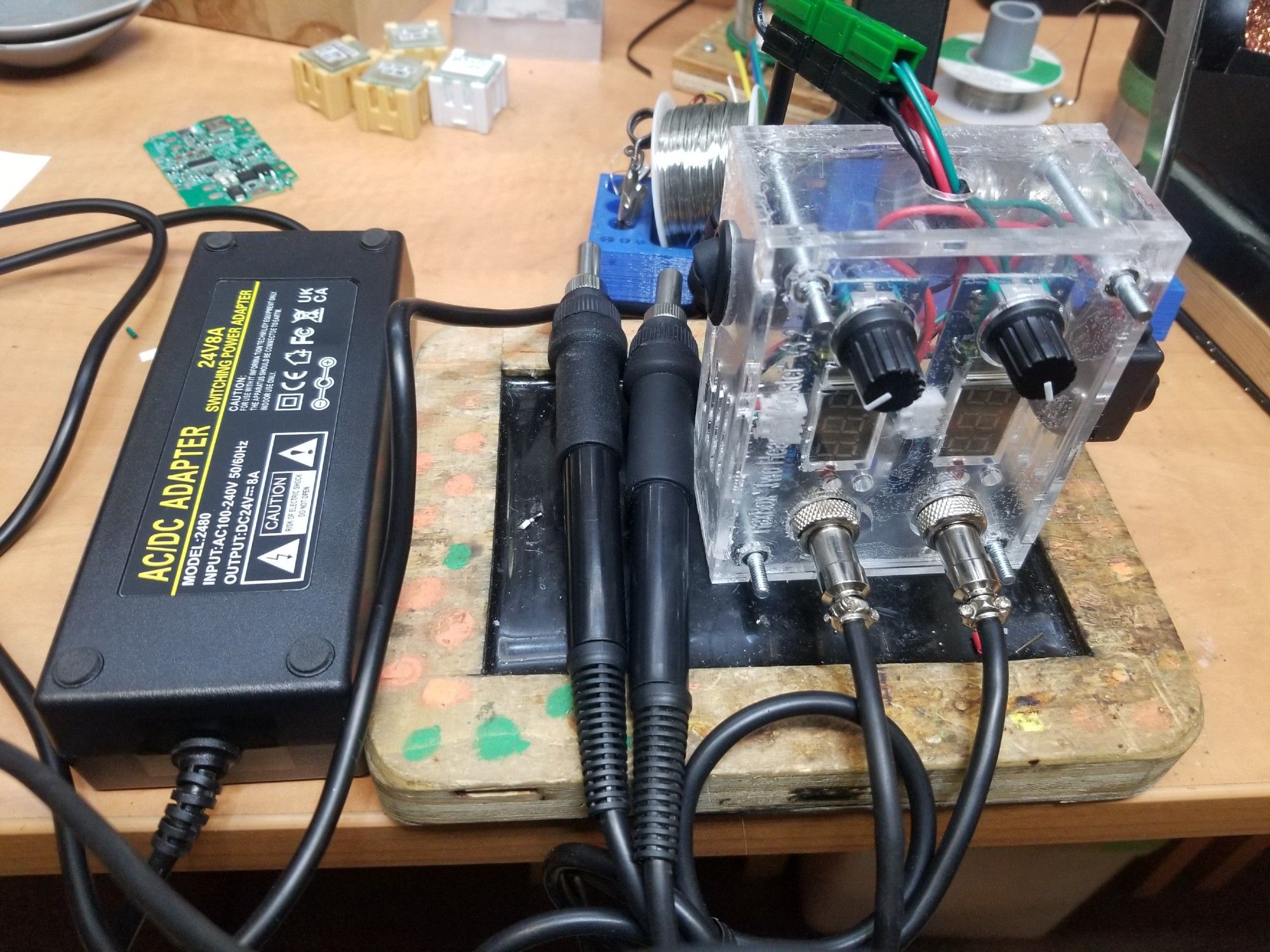 A homemade T12 soldering station sitting on a black anti-static work pad mounted to a wooden table. Two wand-like black plastic soldering tool grips are cord attached to a pair of nickel connectors on the clear plastic laser cut enclosure for the twin circuits underneath, with LED displays and potentiometer knobs on each. Anderson power-poles in red, black and green connect it to its power supply on the left.