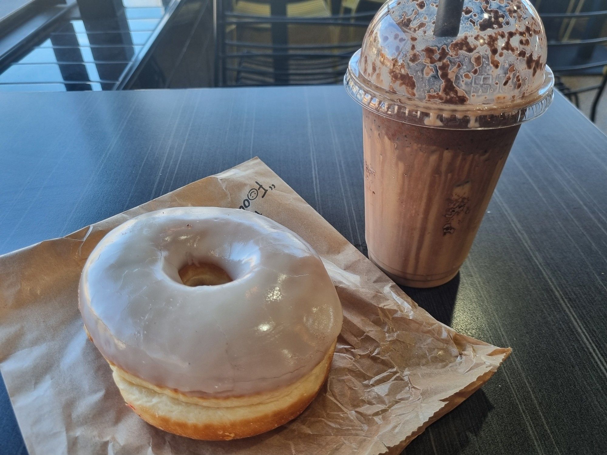 Photograph of a glazed donut and large iced mocha in a transparent plastic cup,  sat on a dark wood table in a petrol station.