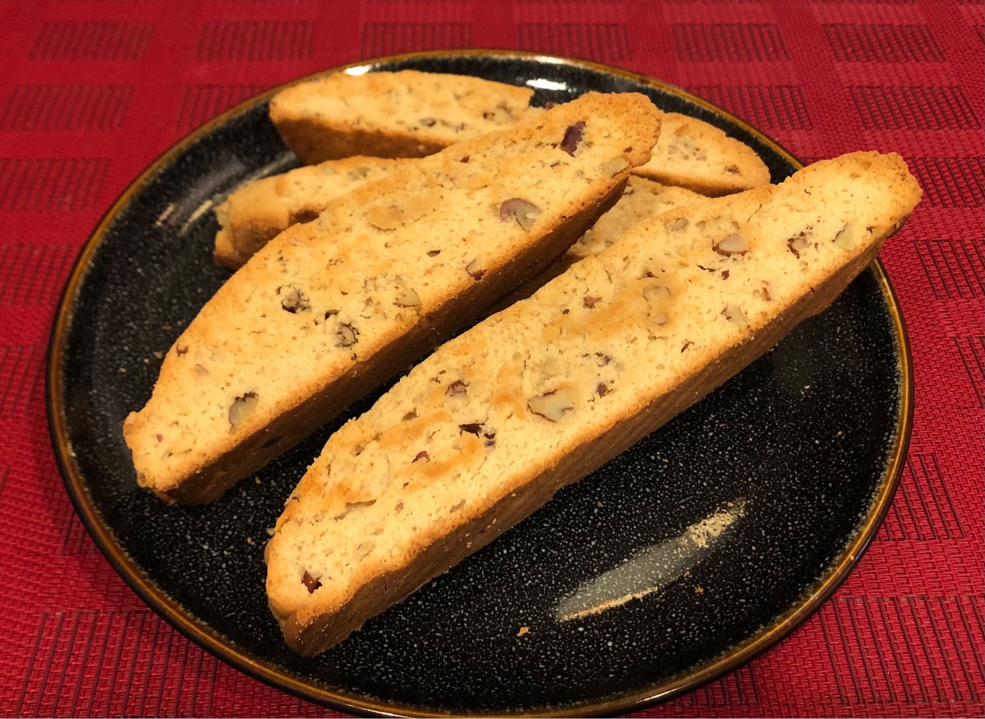 Four pieces of Maple Pecan Biscotti, thick sliced and baked golden brown, on a black plate, on a red placemat. You can see a bunch of chopped pecans in there.