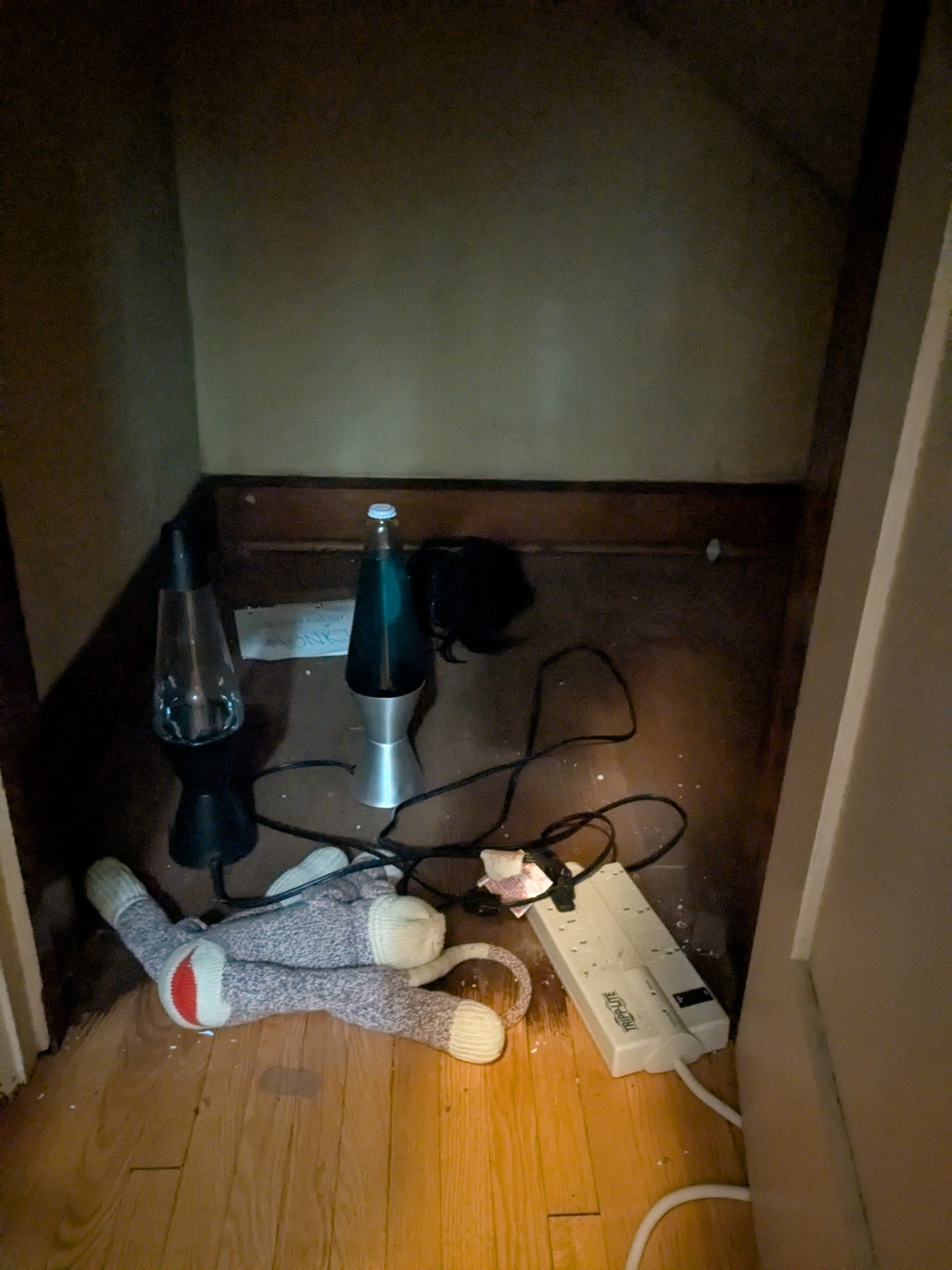 Photo of a small cubby behind a closet, with the low door open to reveal a stuffed monkey, two lava lamps, an extension cord, an old Dora the Explorer wig, and a sign: Organization of MONKI with a fierce monkey face logo drawn on it. It's probably better if you ask no more questions than I did. 