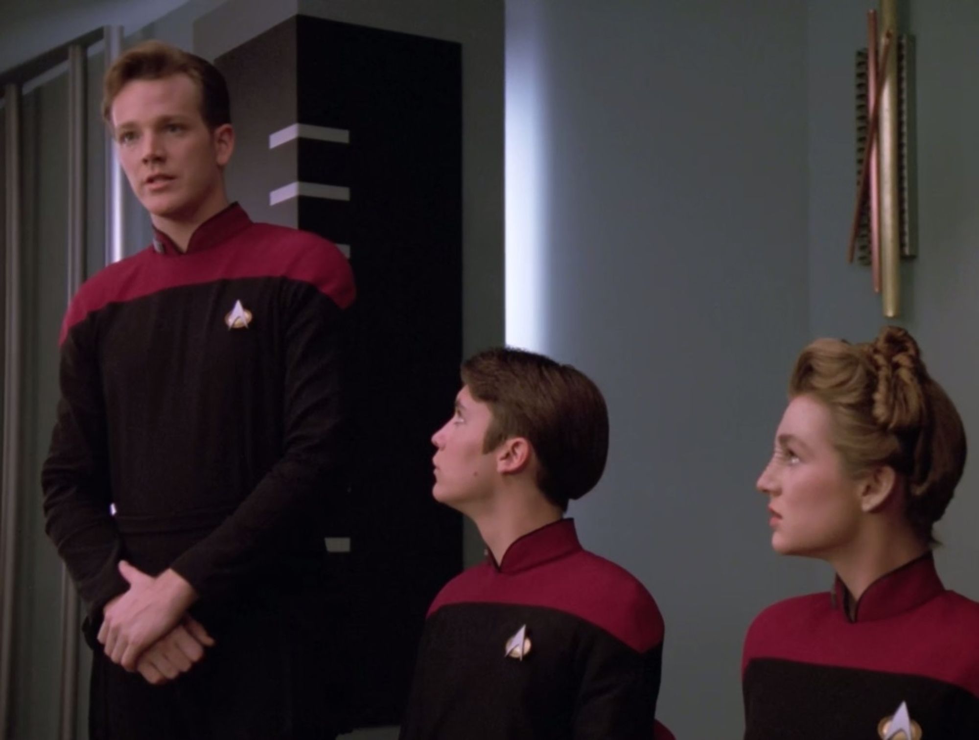 Cadet Locarno stands at his hearing, with his hands folded in front of him. Wesley Crusher and Sito Jaxa sit beside him, watching him with concern.