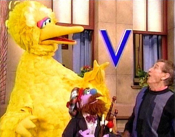 In a scene from Sesame Street, Bob, Big Bird, and a bagpipe player stare up in wonder at a floating letter V.