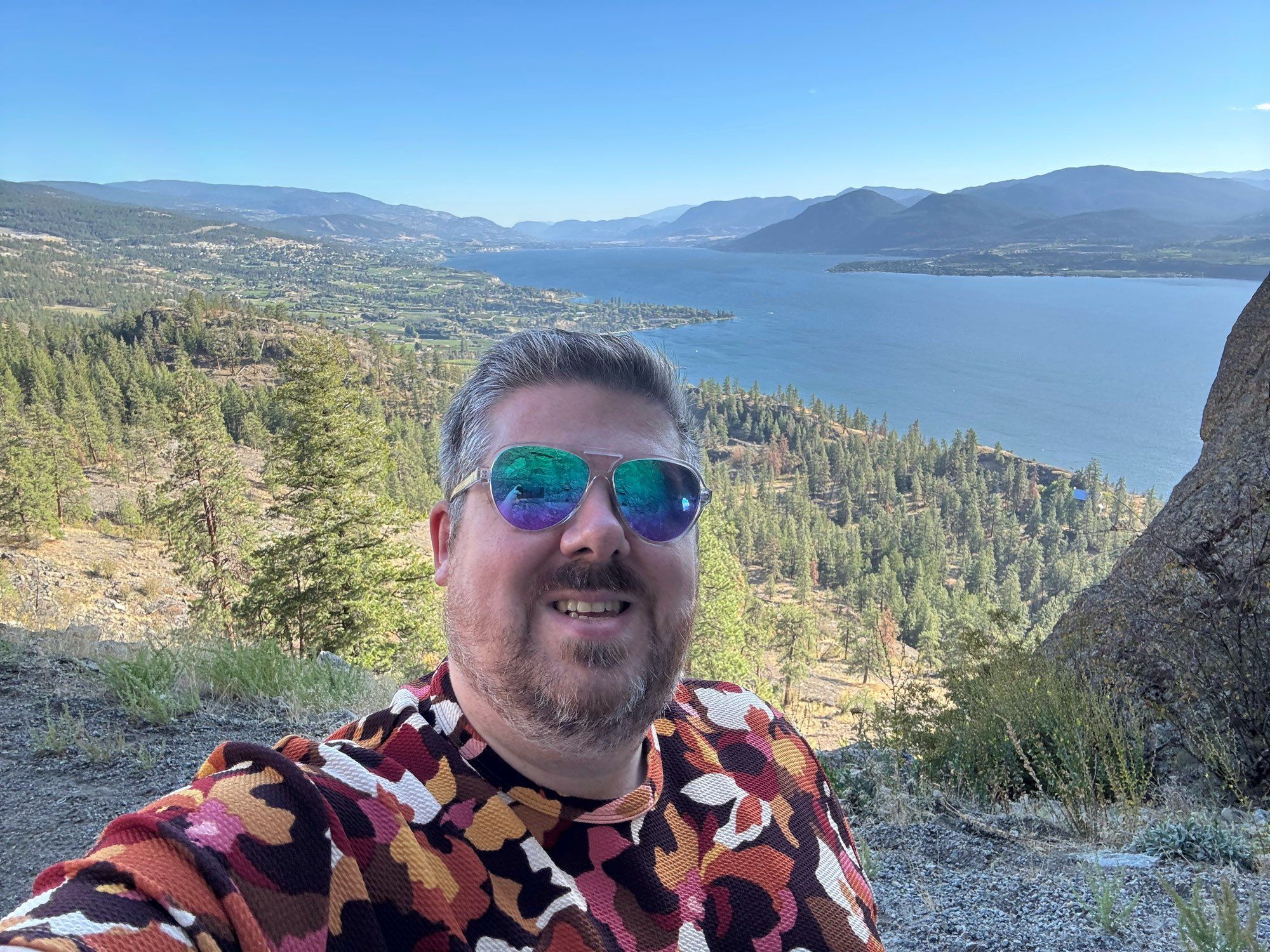 Selfie of a bearded guy wearing sunglasses and a colourful shirt with Okanagan Lake and the communities of Naramata, Penticton and Summerland in the background