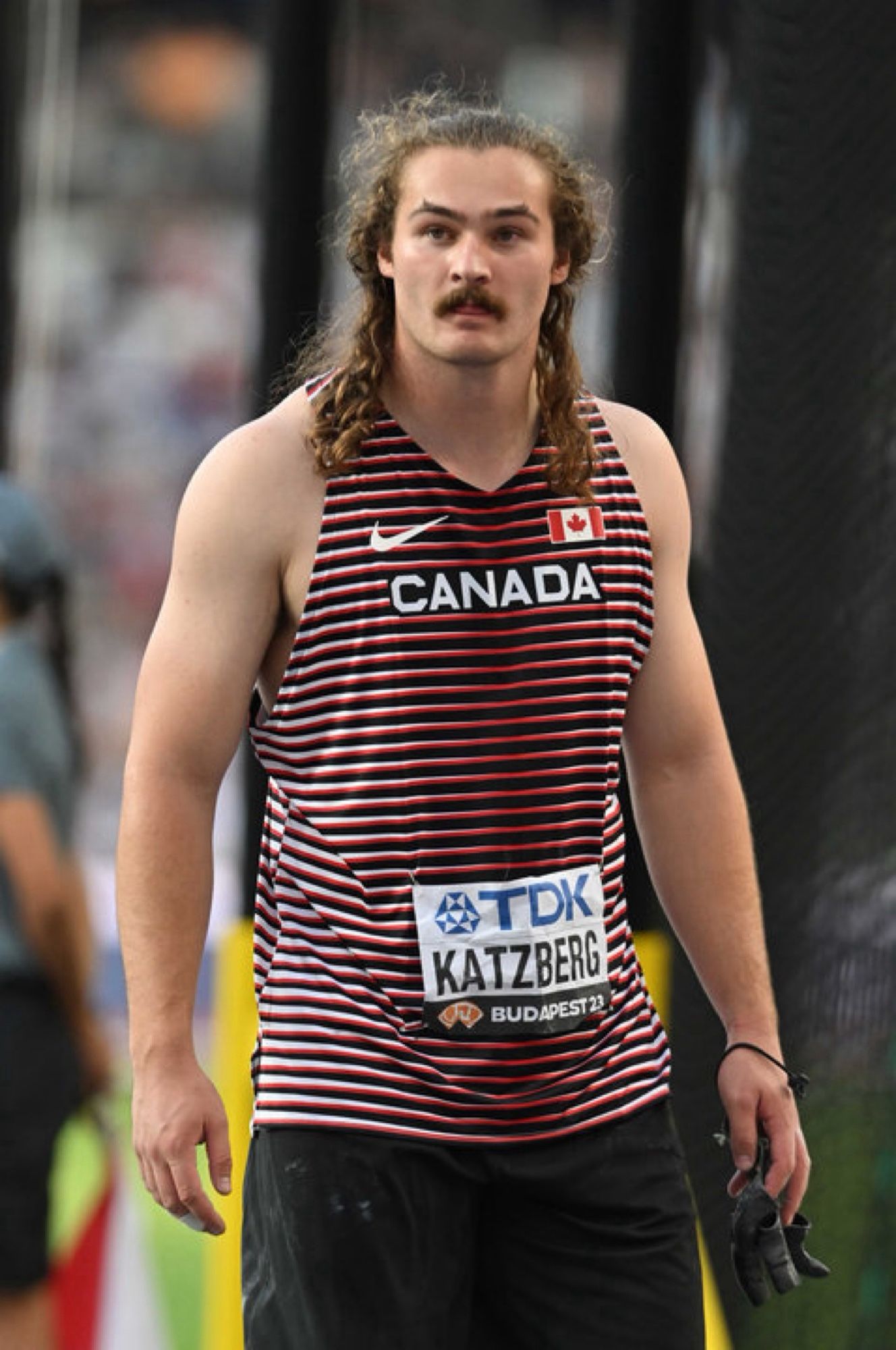 A pretty regular looking tall guy with ridiculous long hair, pointy eyebrows and big mustache. Wearing a Canada singlet.