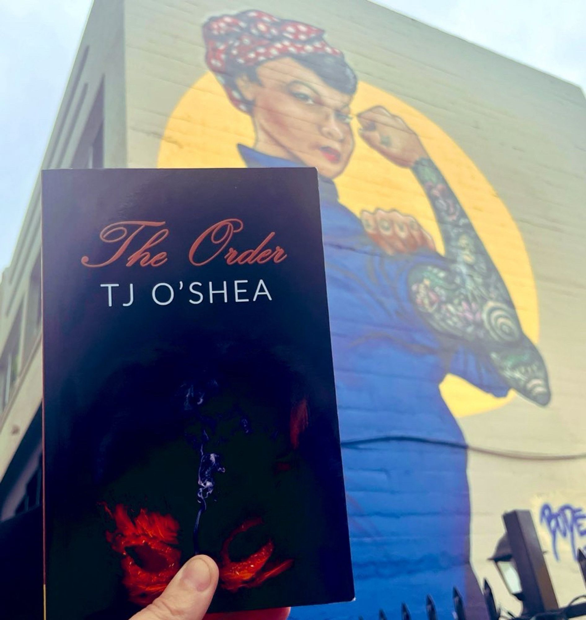 A paperback copy of The Order by TJ O’Shea held in front of a wall mural at Sacred Rose Tattoo in Berkeley.