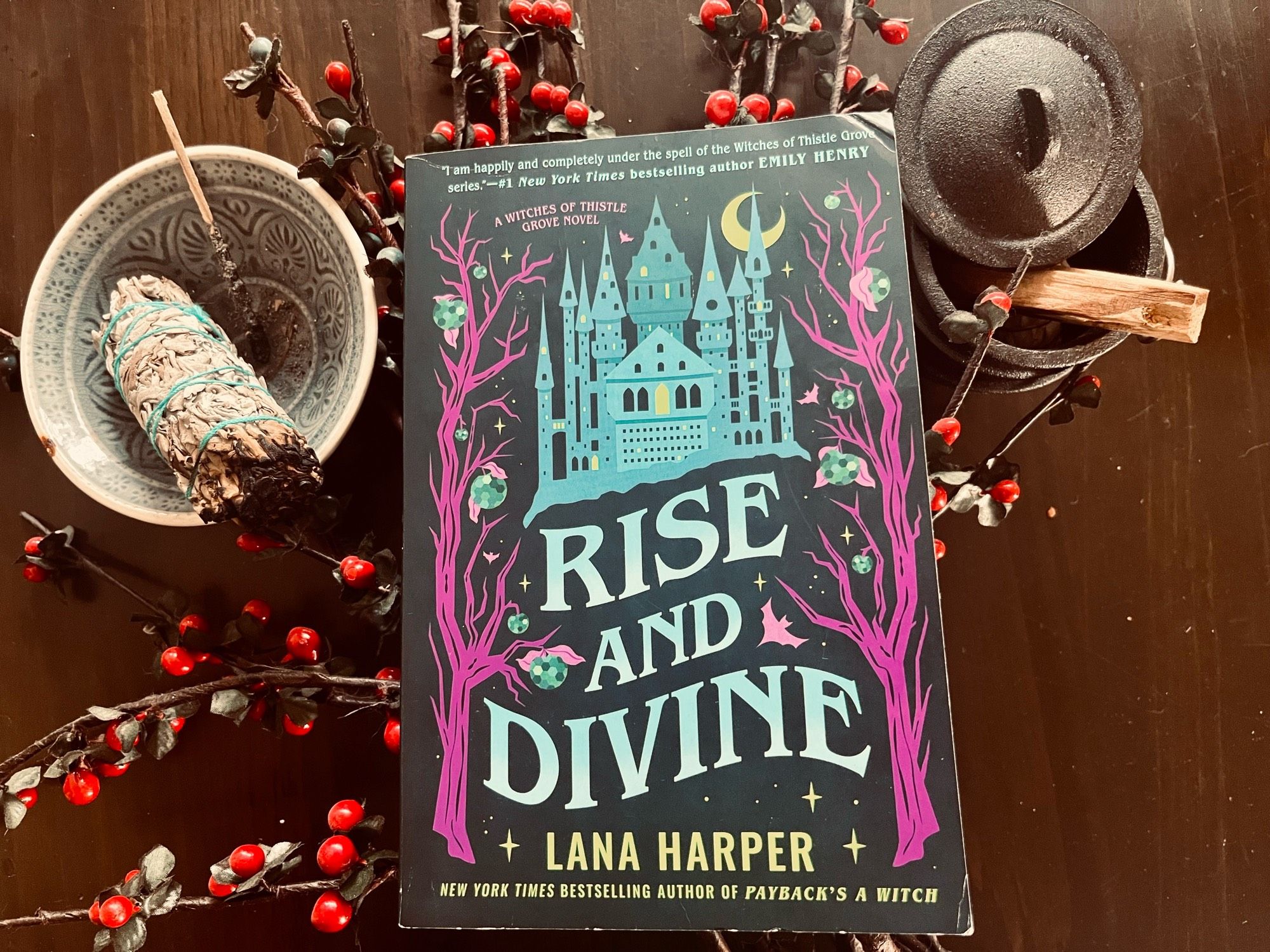 A paperback copy of Rise and Divine by Lana Harper against a bouquet of red berries, a sage smudge stick and a small cauldron.