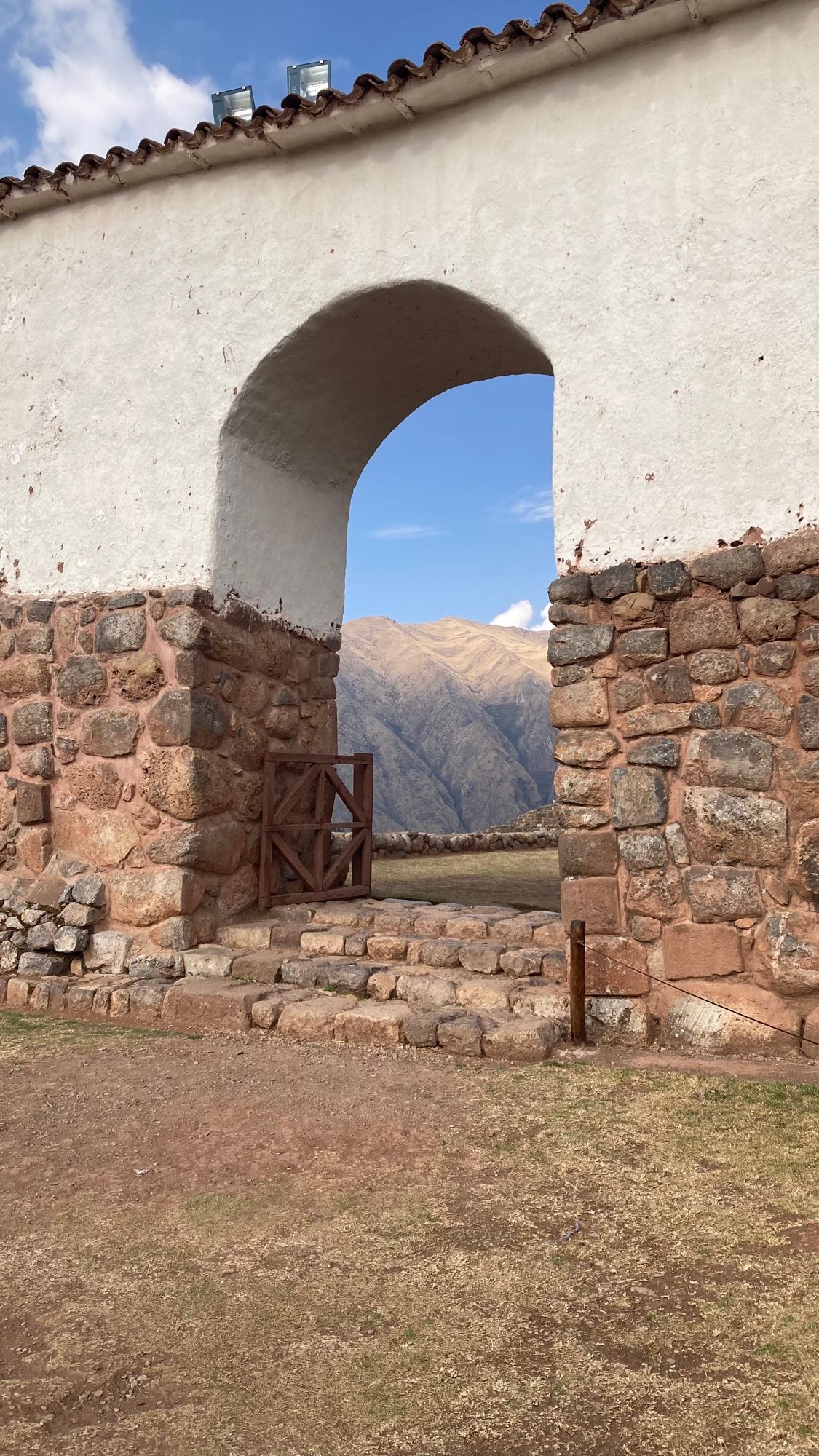 Portal arredondado (com base de pedra e parte de cima brava) da igreja de Chinchero, enquadrando montanhas verdes e bege e o céu azul.