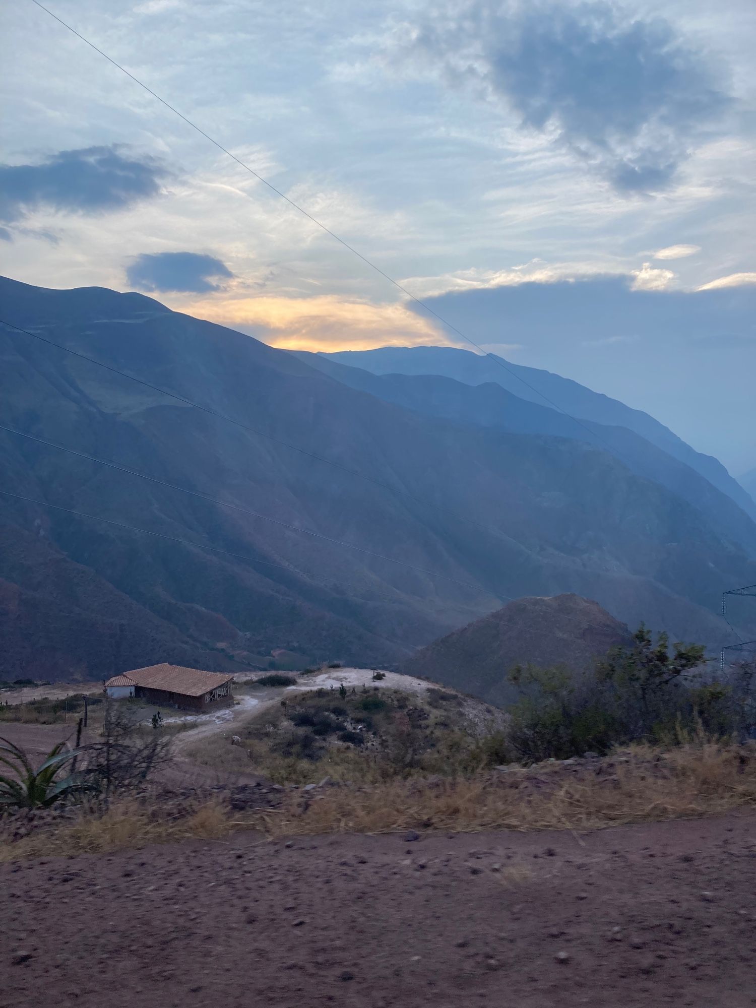 Montanhas verde-escuras contra um céu que começa a escurecer. Diante delas se vê um chão marrom com vegetação esparsa e uma casa ao longe