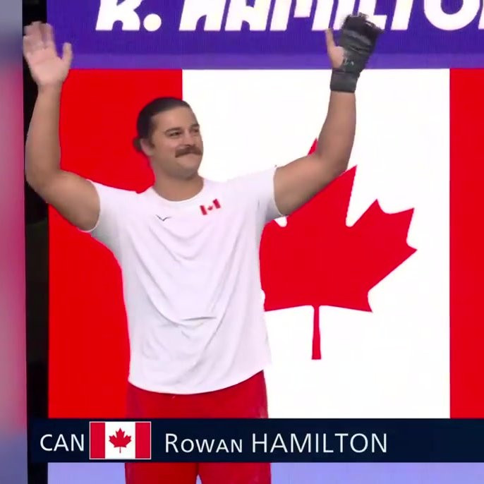 photo of canadian olympic hammer thrower Rowan Hamilton, a white man with a moustache and long hair tied up in a bun, wearing a white t shirt with a small canadian flag on it, standing in front of a canadian flag with his hands held up in the air