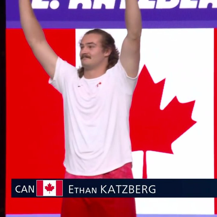 photo of canadian olympic hammer thrower Ethan Katzberg, a white man with a moustache and long hair, wearing a white t shirt with a small canadian flag on it, standing in front of a canadian flag with his hands held up in the air