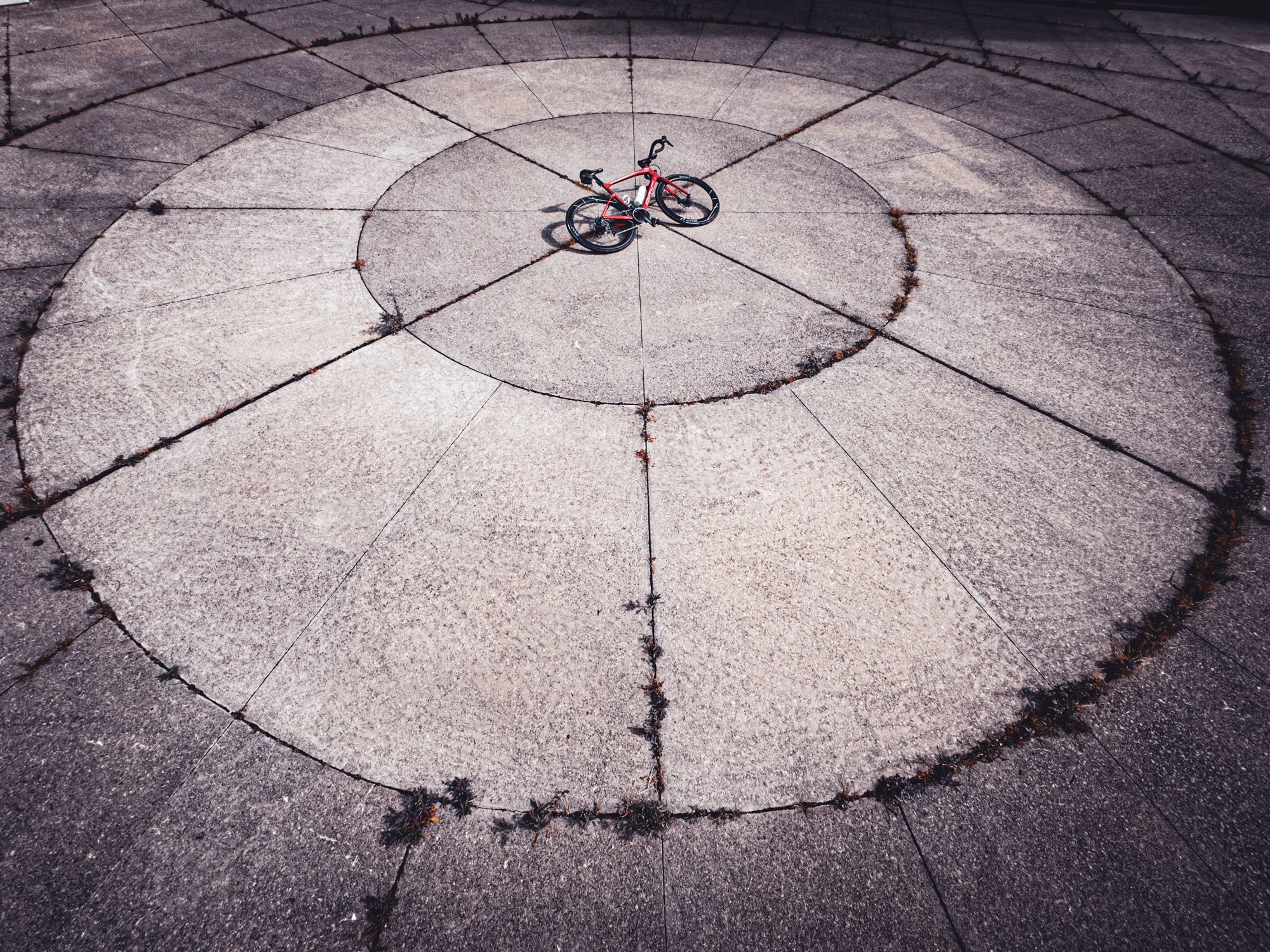 Photo of a red BMC roadmachine, road bike lying on its side in the centre of a circular concrete surface made of pie shaped pieces of concrete. There are three rings of concrete slabs and the bike is sitting right in the center. There are weeds growing between the cracks of some of the concrete slabs.