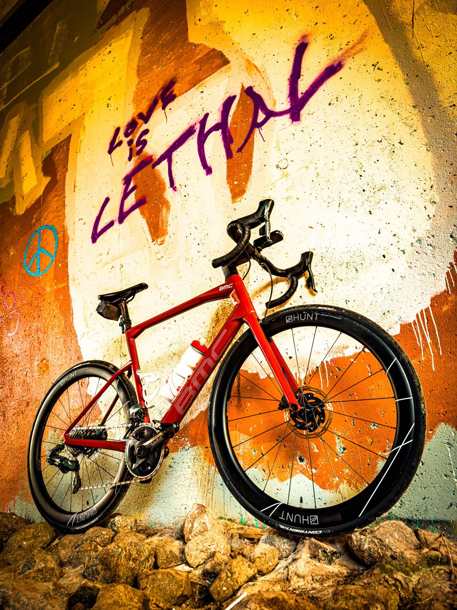 Photo of a red BMC roadmachine load bike standing next to the concrete wall of an underpass. The wall is covered in graffiti with large swats of orange and white and yellow paint. Above the bike are the words “love is lethal”. And next to that is a little blue peace sign. The bike is standing on a bed of rocks.