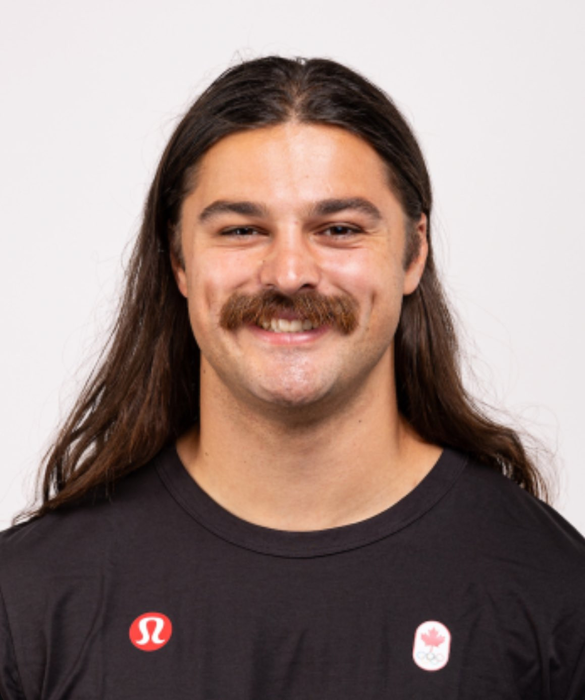 photo of canadian olympic hammer thrower Rowan Hamilton, a white man with a moustache and long hair, wearing a black t shirt with a small canadian olympic logo on it and a lululemon logo, smiling