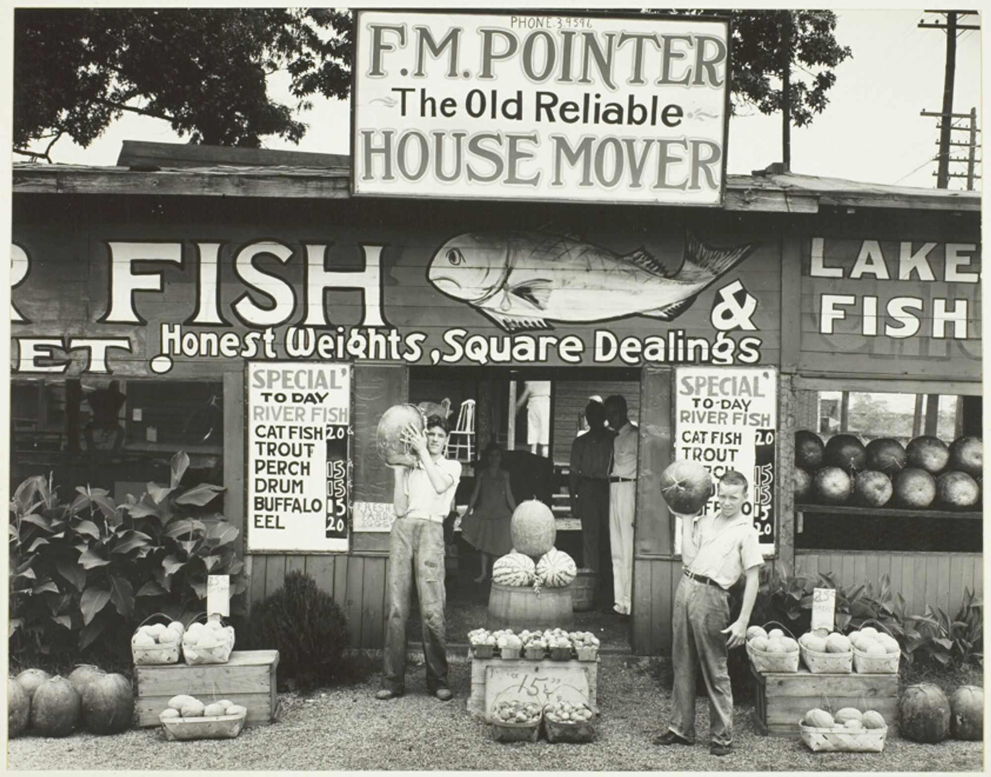 This is a black & white photograph of a roadside establishment, which touts its “Honest Weights, Square Dealings.” An assortment of fruits, melons & plants are displayed in-and-around an open-air market. Signs posted on the building highlight the variety of fresh fish for sale. A large sign posted above the entrance advertises the services of “F.M. Pointer – The Old Reliable House Mover.”
     “Roadside Stand Near Birmingham” 1936 by Walker Evans (American, 1903–1975)
     “[Walker Evans’] photographs of roadside architecture, rural churches, small-town barbers, & cemeteries reveal a deep respect for the neglected traditions of the common man and secured his reputation as America’s preeminent documentarian. From their first appearance in magazines & books in the late 1930s, these direct, iconic images entered the public’s collective consciousness & are now deeply embedded in the nation’s shared visual history of the Depression.” – Dept of Photographs, The Metropolitan Museum of Art