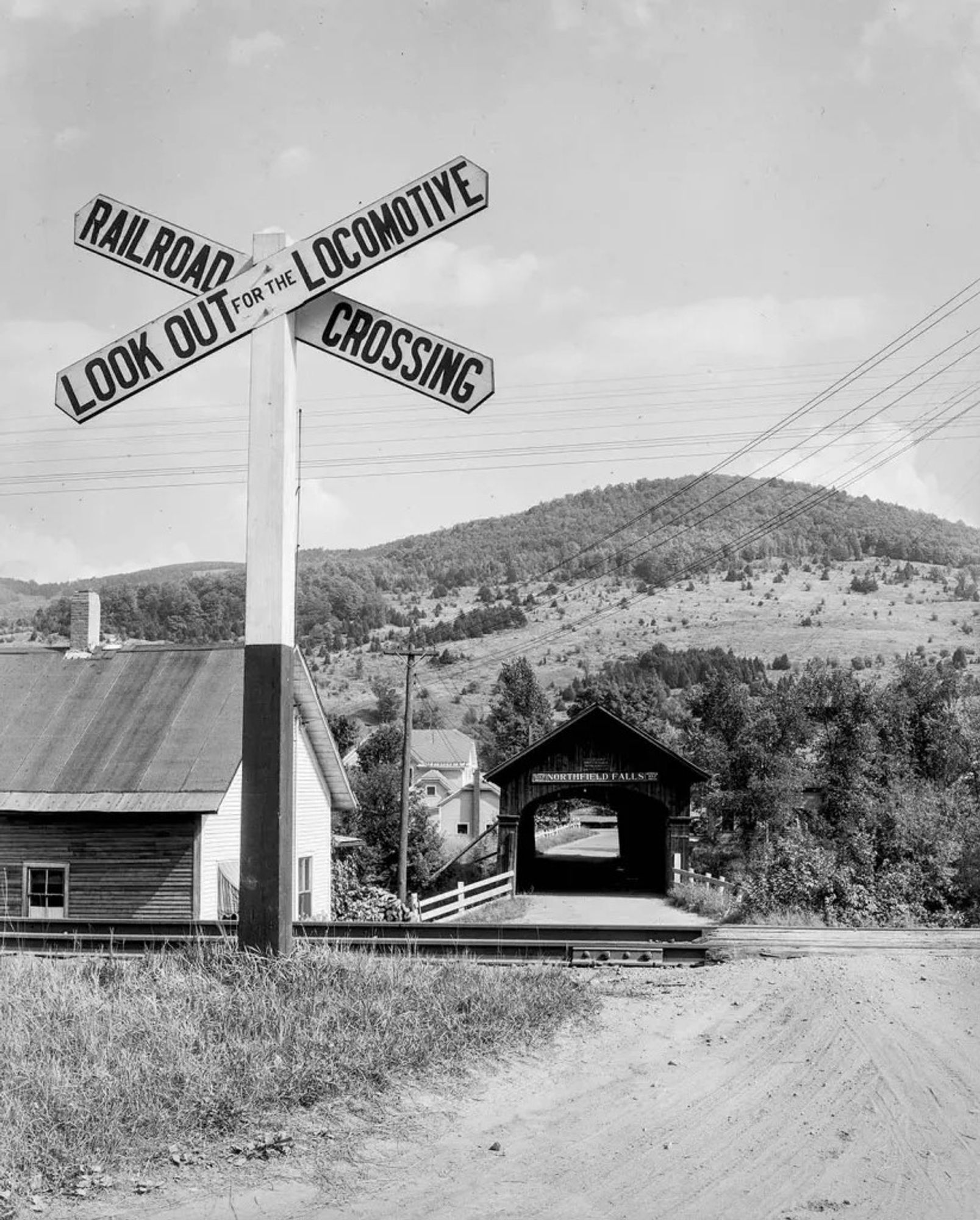 This is a black & white photograph. It features a railroad crossing sign that is situated to the left of a dirt road. The sign does not have the flashing red lights & crossing gates that, these days, usually block the roadway as a train approaches. Beyond the crossing is one of the four covered bridges that Northfield Falls, VT is known for. This one crosses the Dog River on the west side of the village. 
     Source: Farm Security Administration - Office of War Information Photograph Collection at the Library of Congress