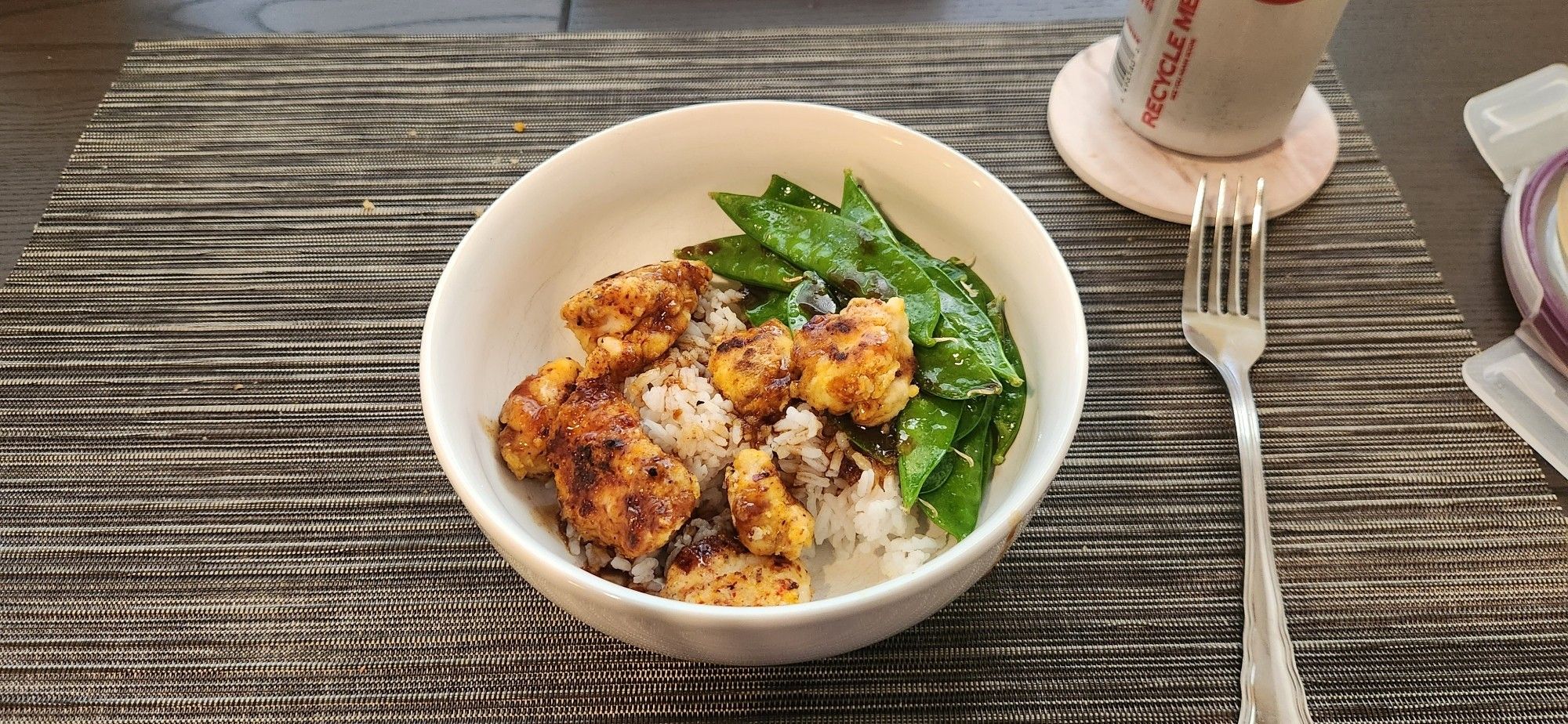 A white bowl on a dark table with browned, breaded chicken and bright green snow peas and a thick dark sauce drizzled over. A bed of rice peeks through from underneath