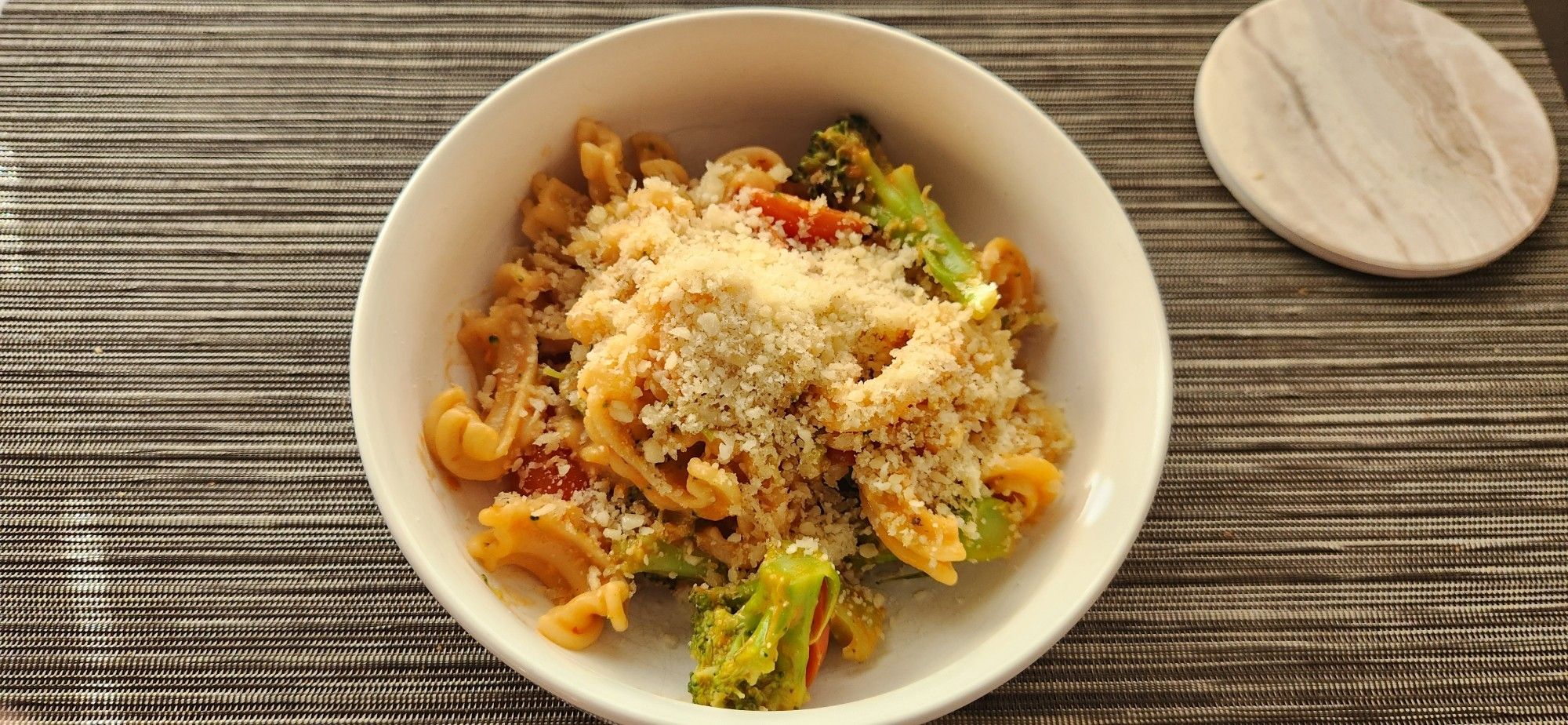 A white bowl on a dark table inside is a serving of cascatelli pasta with broccoli and a tomato sauce, topped with a generous supply of breadcrumbs and parmesan cheese