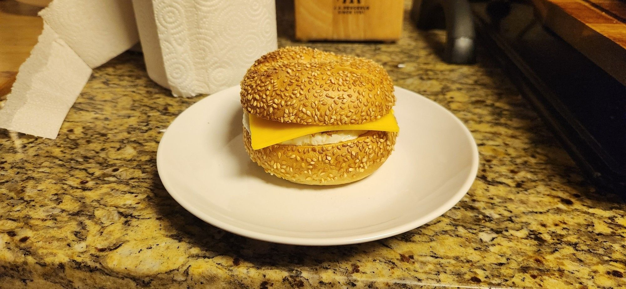 A toasted sesame bagel breakfast sandwich on a white plate. A bit of egg and the corners of a slice of cheese peek out from the middle