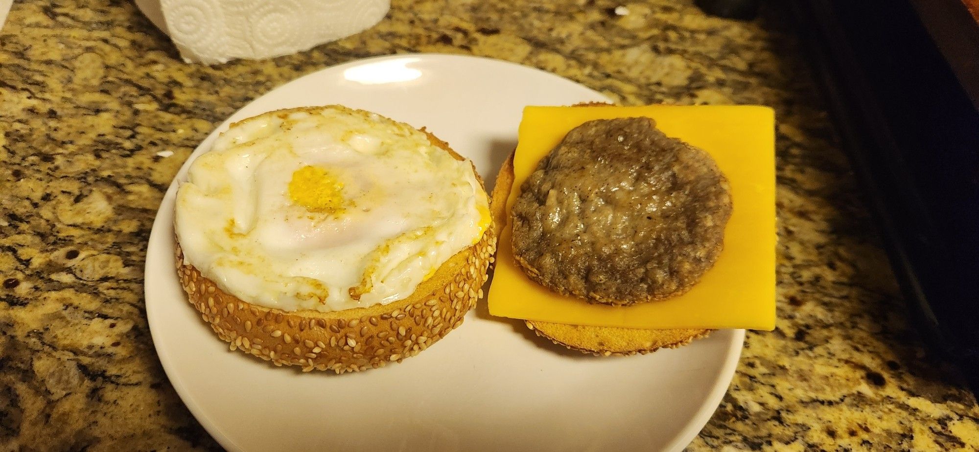 A toasted sesame seed bagel on a white plate. The left half has a fried egg, and the right half has a small sausage patty on a larger square of yellow cheddar cheese