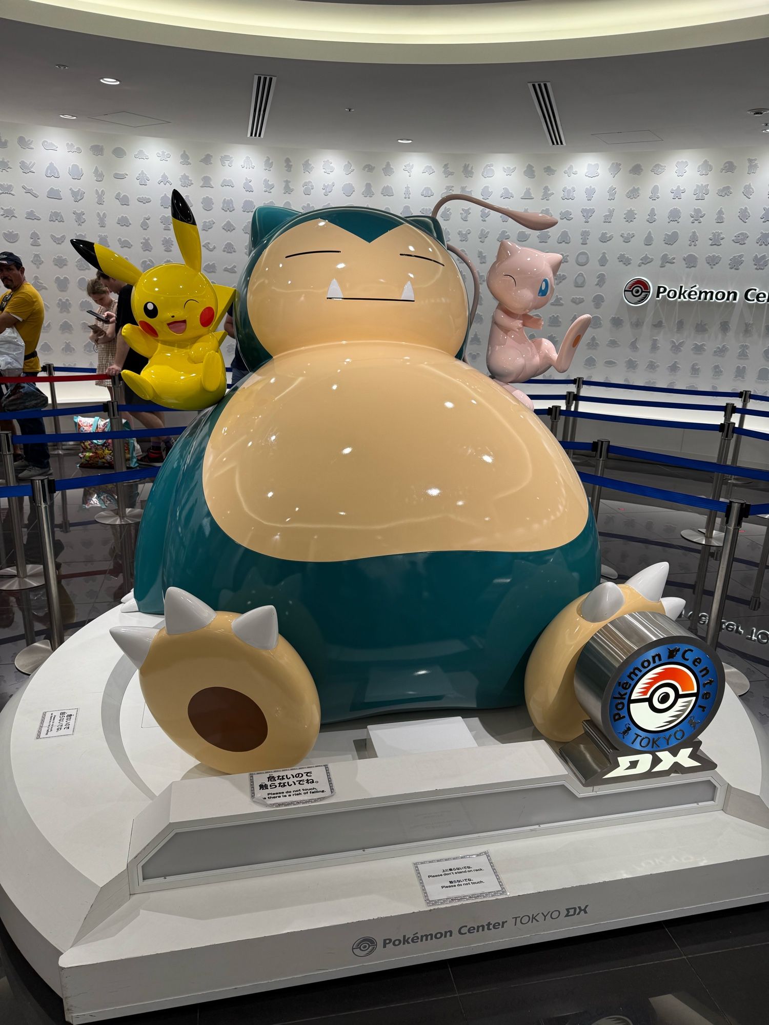 Photo of a giant Snorlax statute. Pikachu is on its right shoulder, Mew is on its left shoulder. In front of its left foot is the image of a pokeball over the letters "DX"