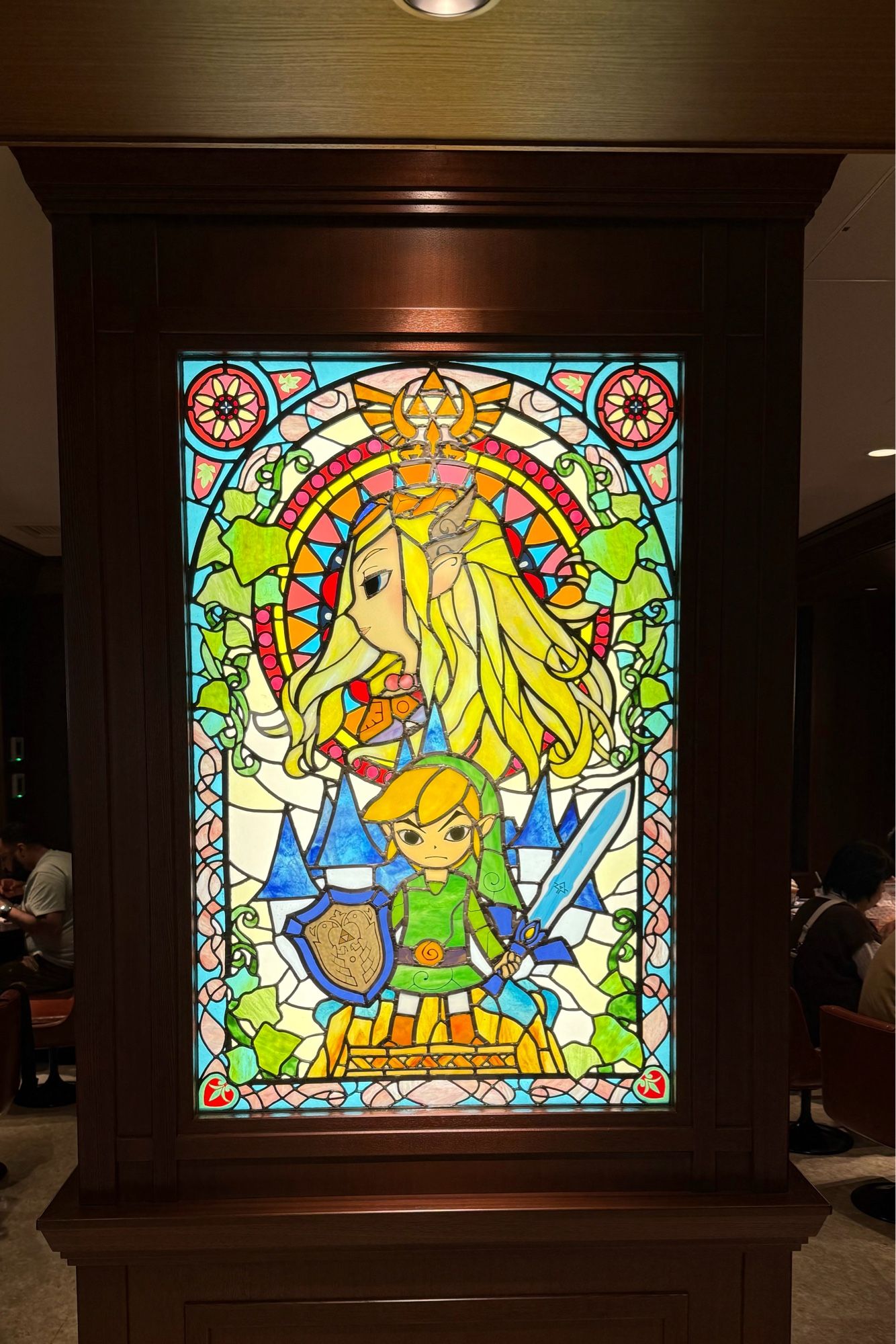 Photo of stained glass artwork, featuring the Legend of Zelda. There is a Triforce in the top middle, a profile/side image of Princess Zelda below it, and an image of Link facing forward below here. Link is holding the Master Sword and the Hyrulean Shield.