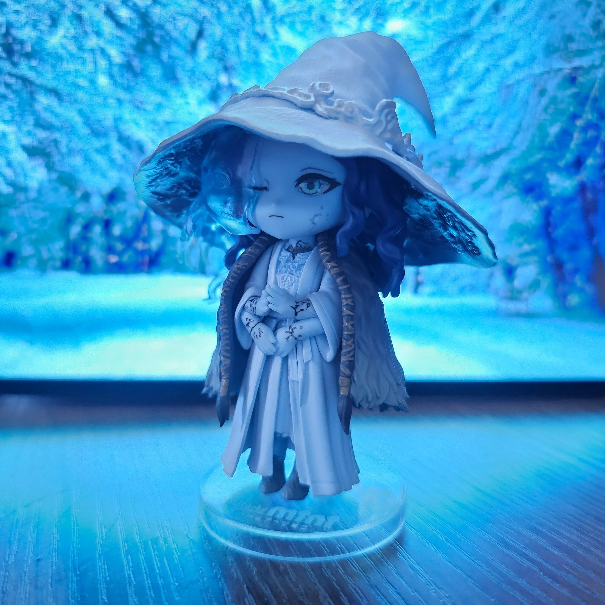 A photo of the new Ranni the Witch figure I got -- Bandai Spirits Figuarts Mini 156. It's very cute, and I'm happy to have some official Ranni merch. I found a pic of a winter forest and displayed it on the computer monitor behind her, so the photo has a cool blue glow to it.