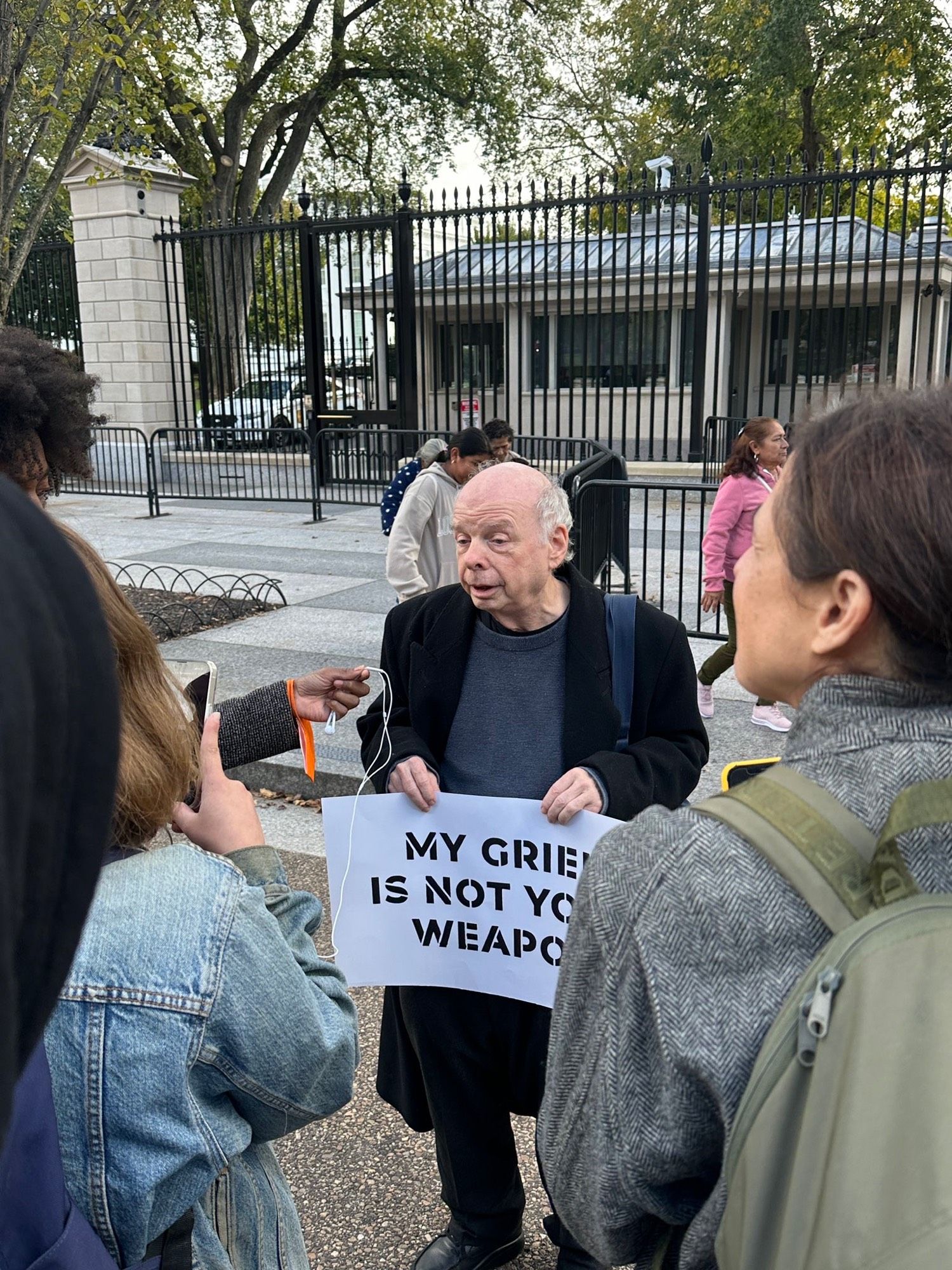 Actor Wallace Shawn, from such classics as The Princess Bride, stands in front of the White House at an If Not Now protest for a cease fire in Gaza/end to the genocide. He holds a white protest sign with black lettering that reads “My grief is not your weapon” as he speaks with reporters