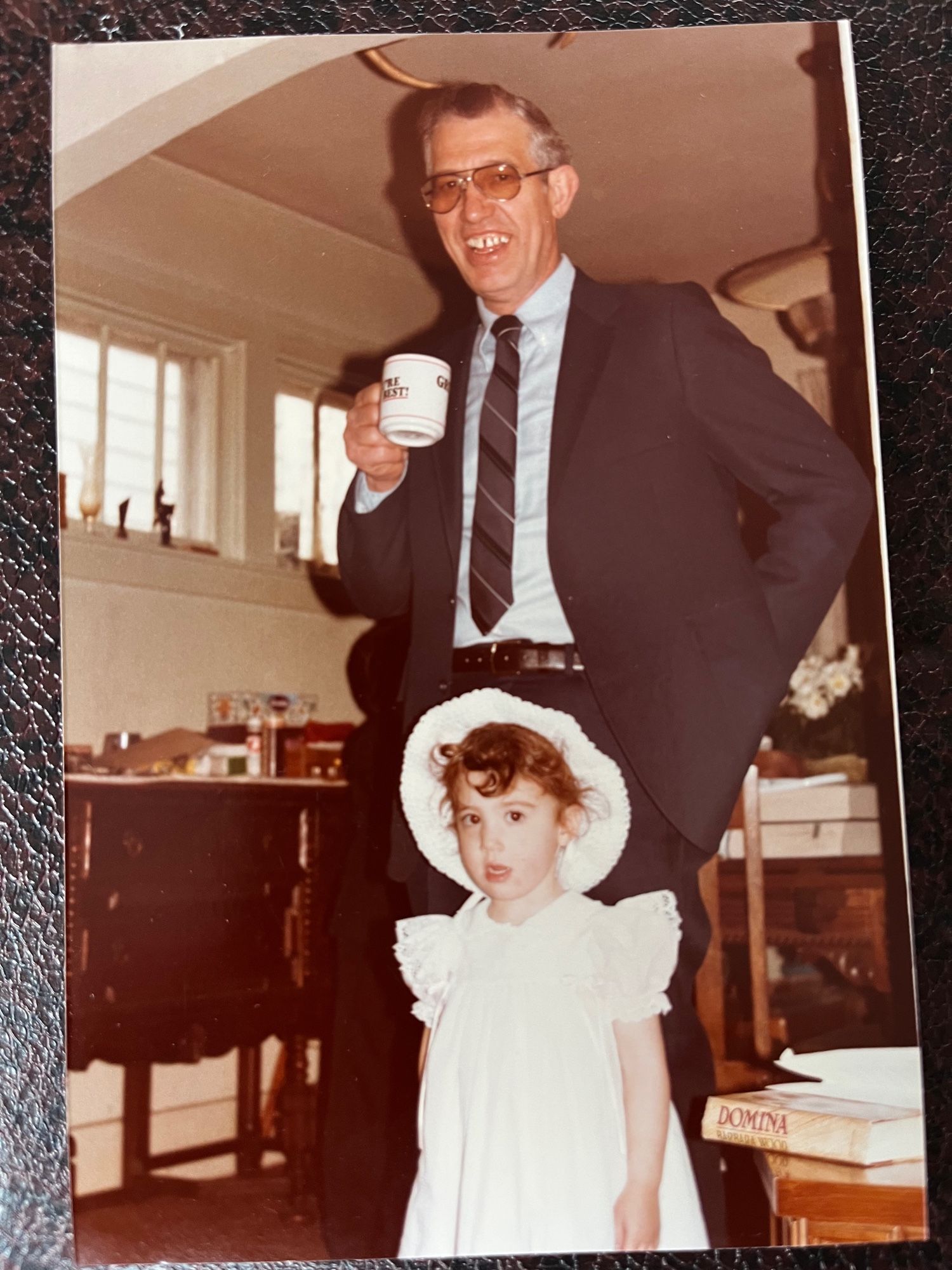 An old photograph of me in my Easter dress and bonnet standing in front of my grandpa who is wearing a navy blue suit. He’s holding a coffee mug and smiling for the camera. I look like I might start crying