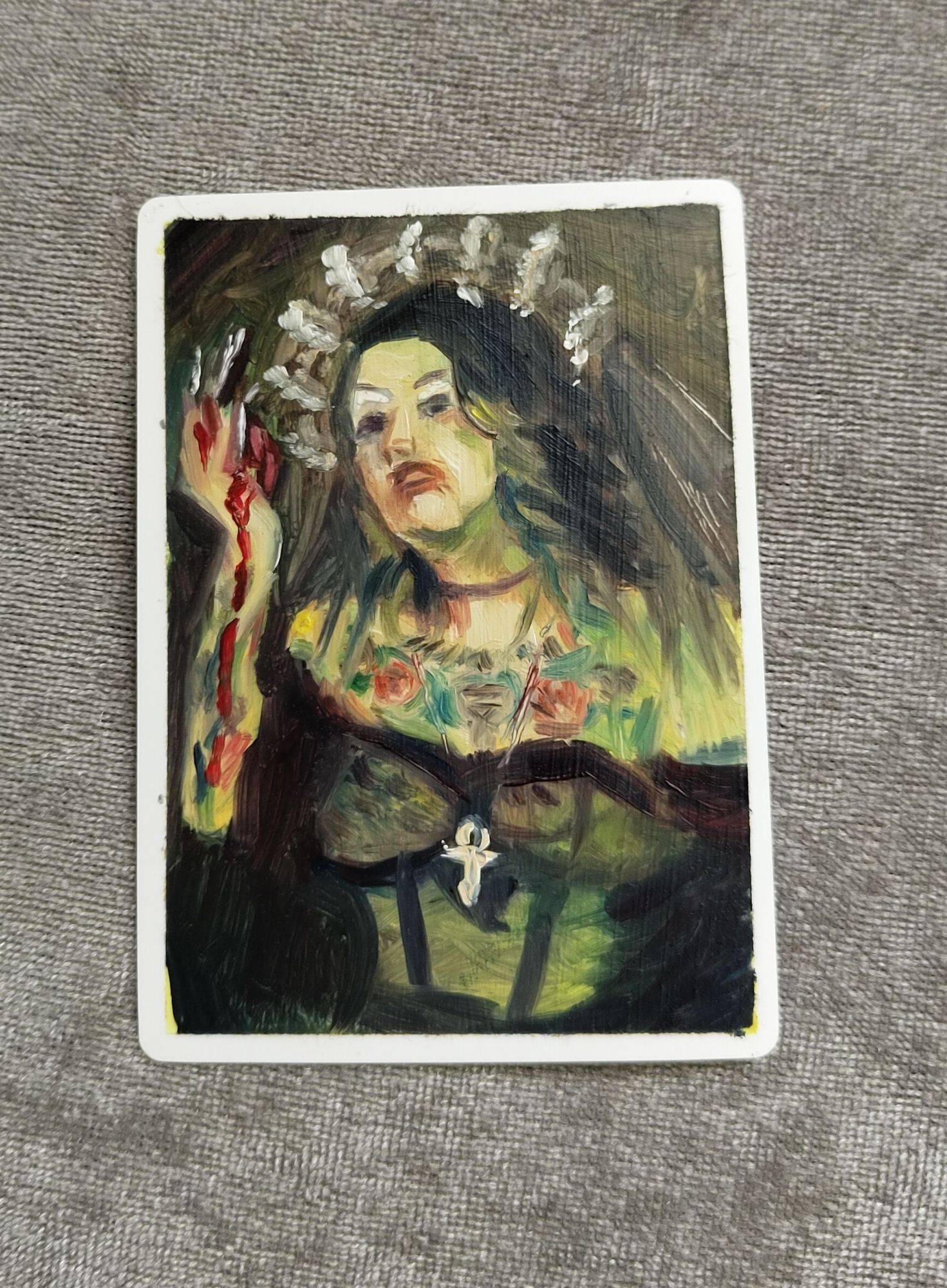 A small oil painting of a long haired, tattooed person with a tiara on, an ankh necklace, holding a heart. The main hue to the skin is green.