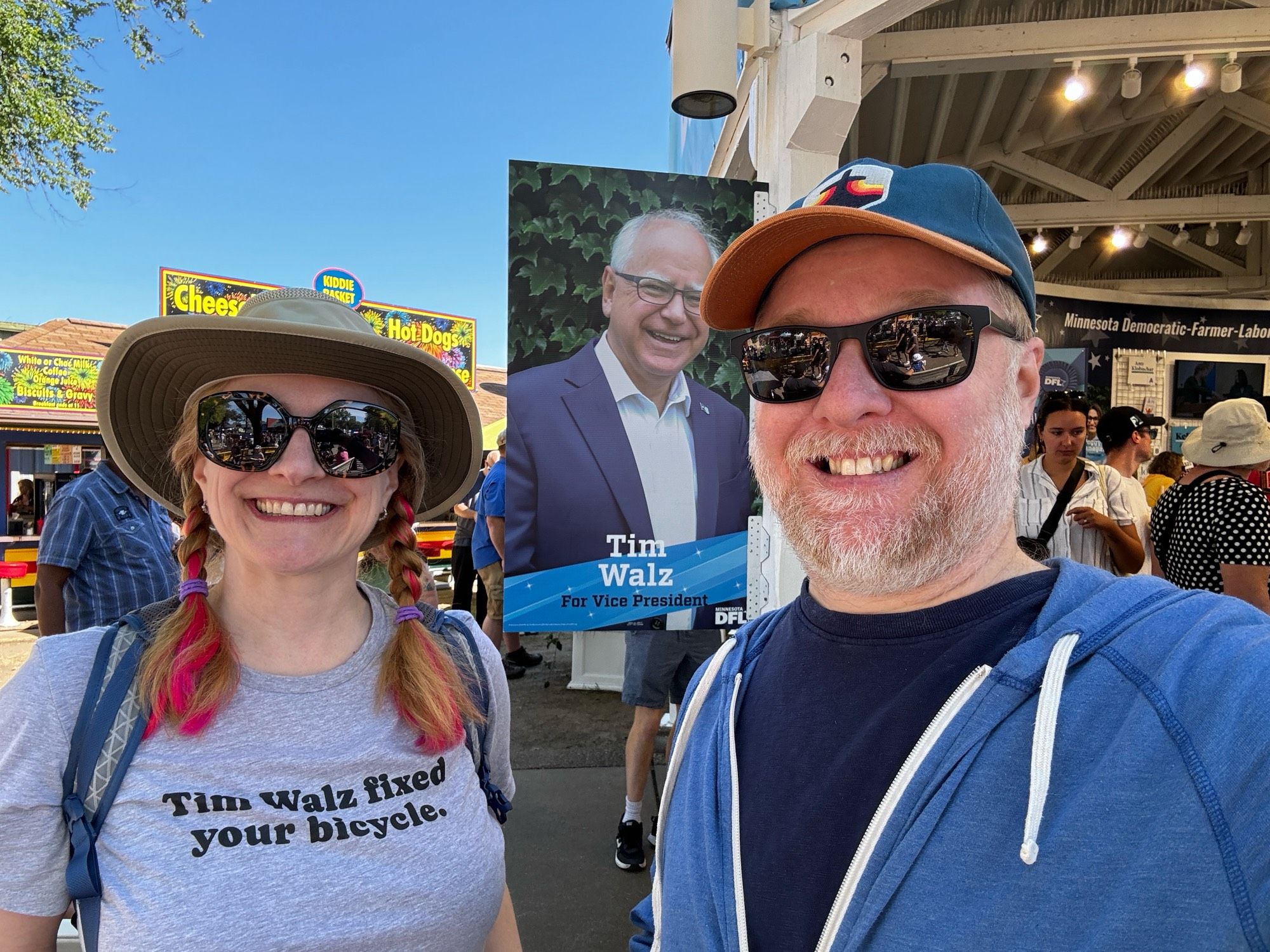 Bridget and Joe at the MN State Fair DFL booth by a Tim Walz sign. Bridget’s shirt says, “Tim Walz fixed your bicycle.”