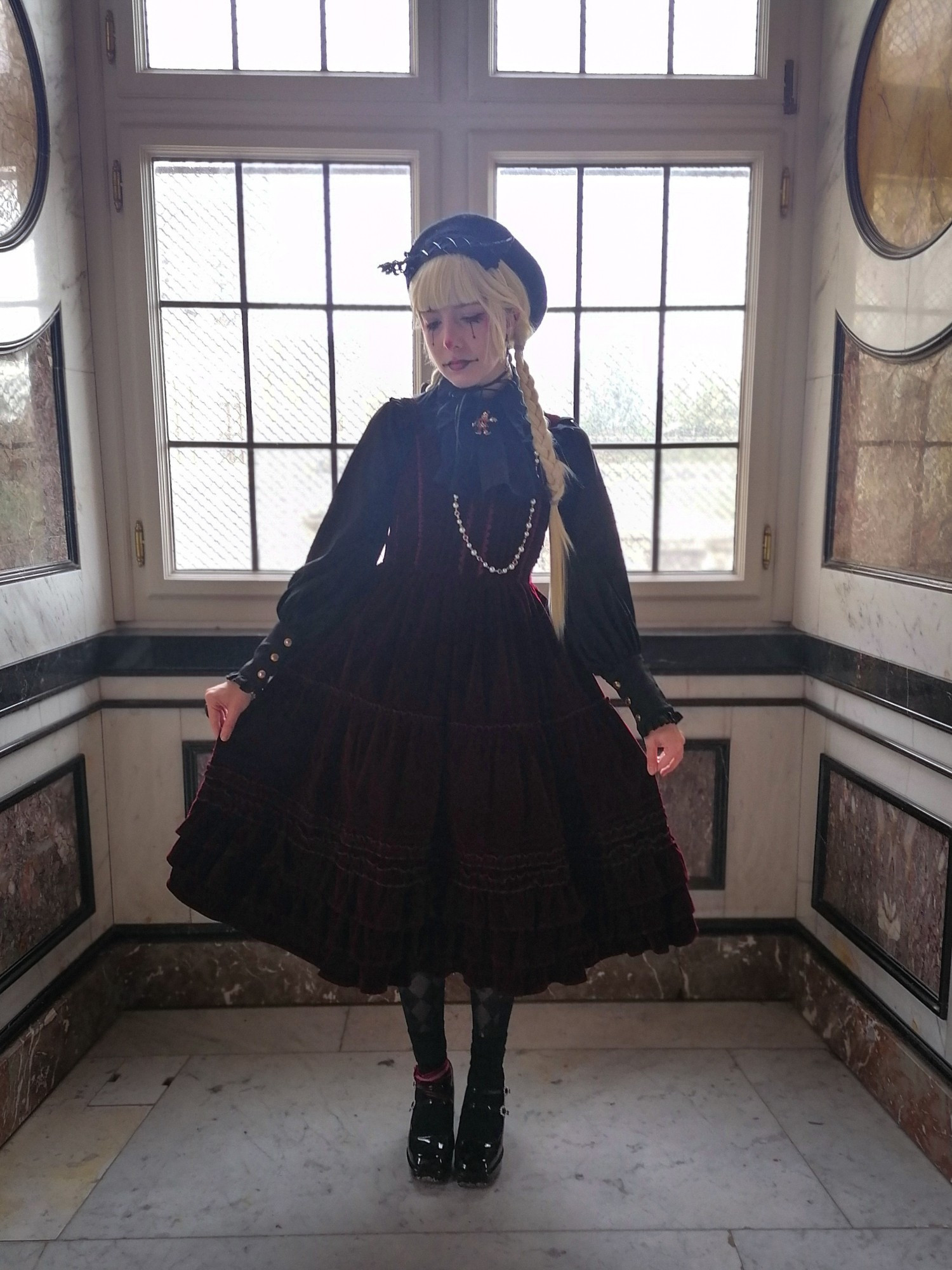 Laney is wearing a lolita fashion coordination, featuring a dark red velvet dress and black accessories. They are wearing a blonde wig, tied into braids, and pierrot makeup. They are surrounded by artful marble flooring and walls. 