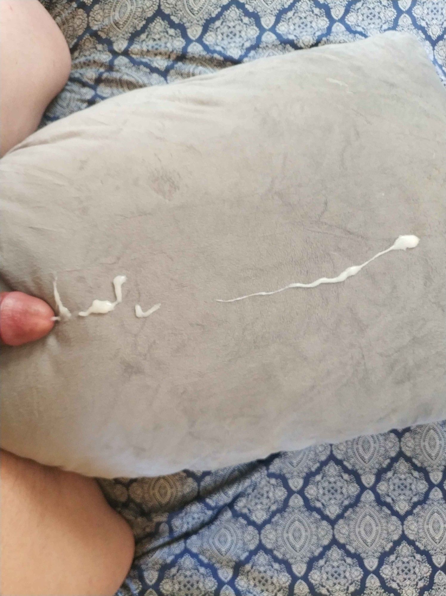 An image of the tip of a penis, with cum splattered across the length of a pillow, and legs on either side of the pillow, the person straddling it.