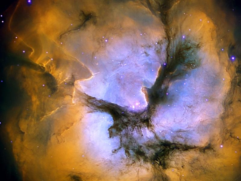 A colorful image of the Trifid Nebula, a star forming region in the Sagittarius constellation. Glowing gas and dust lanes mix together, creating a stunning visual. Three dark dust lanes meet in the center, giving the nebula its name. The image was captured by the 0.8-meter IAC80 telescope on the Canary Islands of Spain.