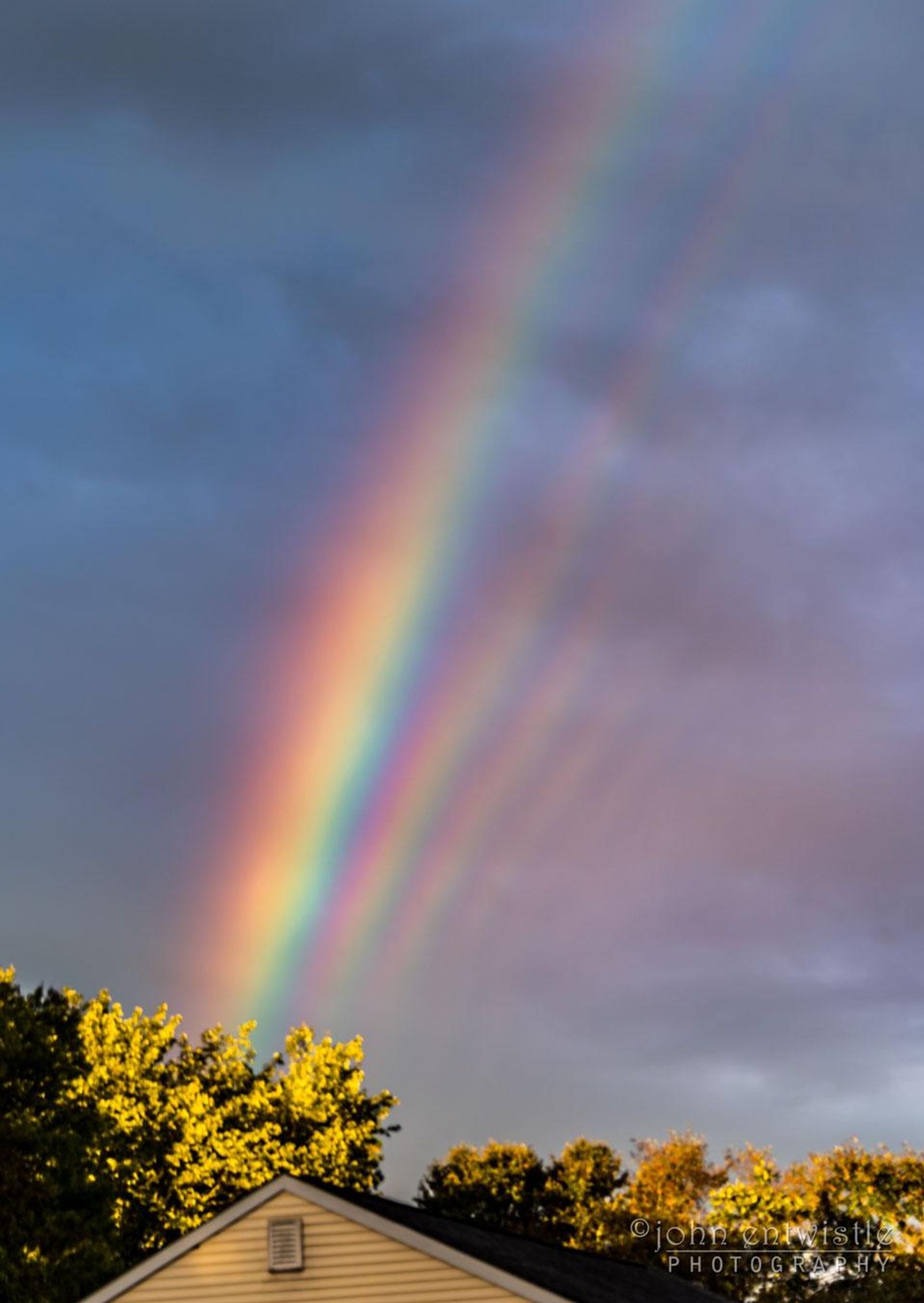 A supernumerary rainbow, a rainbow with multiple parallel bands of color, stretches across a cloudy sky after the remnants of Hurricane Florence passed over the Jersey Shore. The supernumerary bands fade in and out, with at least five captured in this image. The rainbow appears over a line of trees in the foreground and a yellow house.  The photo was taken from the photographer's home in New Jersey. The supernumerary rainbow forms when falling water droplets are all nearly the same size and typically less than a millimeter across, and is a result of light interference.