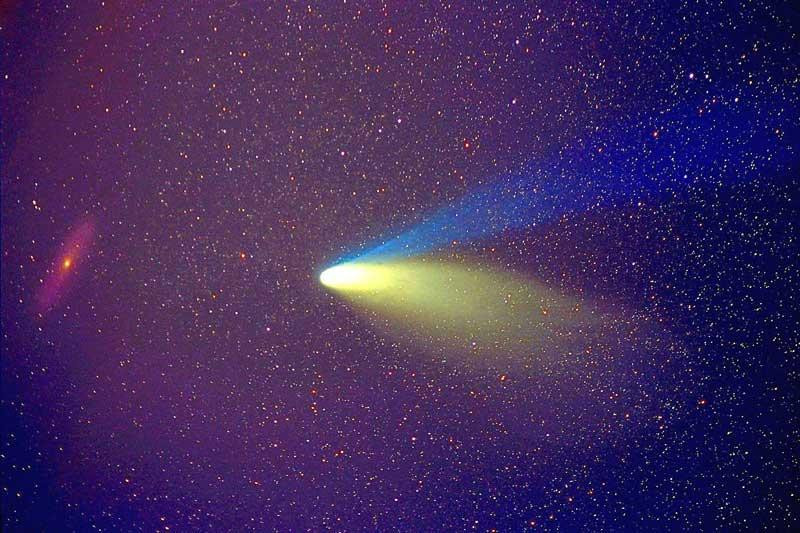 A photograph of Comet Hale-Bopp passing in front of the Andromeda Galaxy, taken during a total lunar eclipse in March 1997. The comet's tail is visible as a blue streak, while the Andromeda Galaxy can be seen as a faint red smudge in the background. The photo was taken during a lunar eclipse to avoid the bright moonlight that would have otherwise obscured the comet and galaxy.