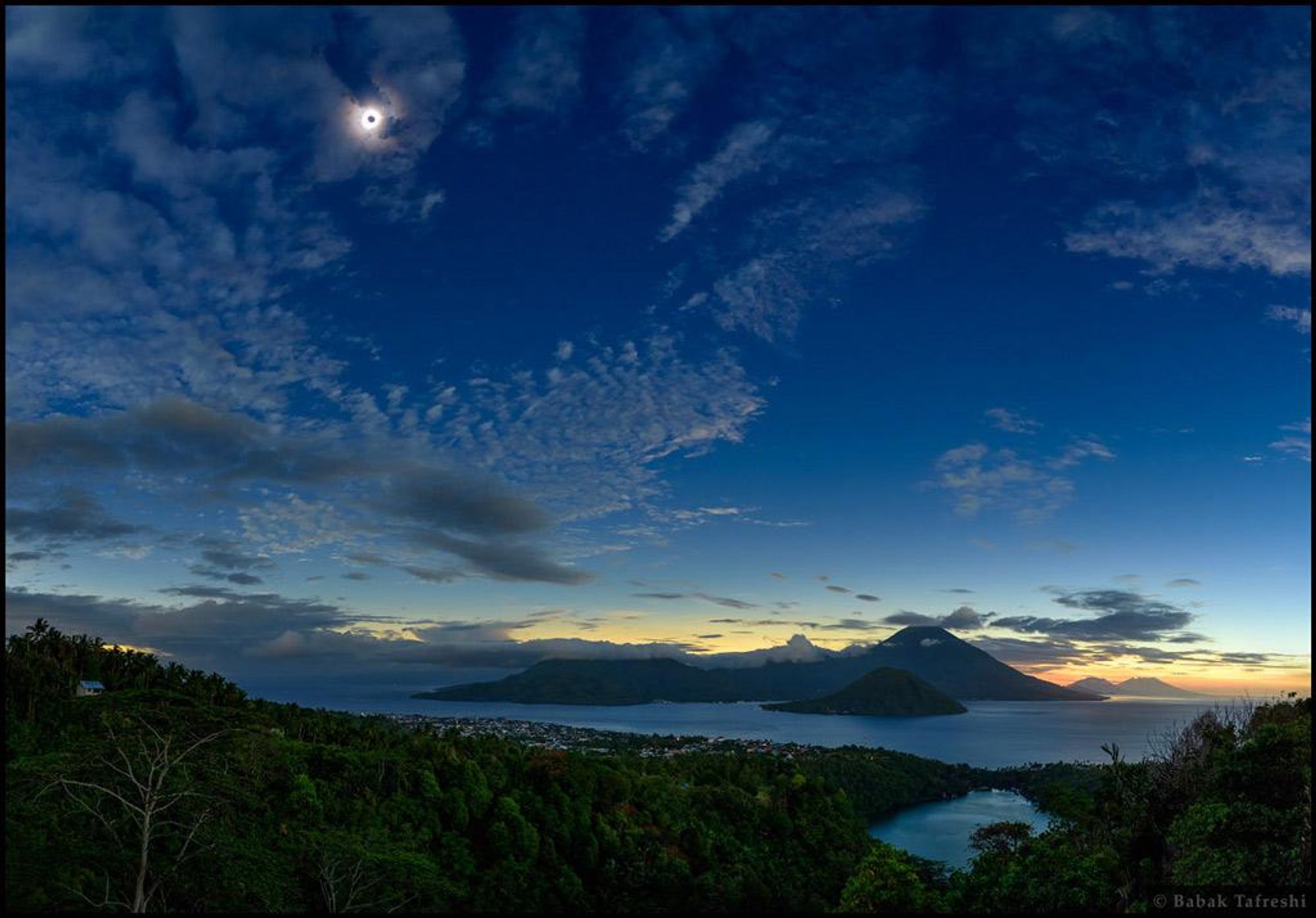 A stunning panoramic view of a total solar eclipse taken from the Indonesian island of Ternate. The dark Sun hangs in the clearing sky over volcanic mountains, with the bright glow of the solar corona shining through thin clouds around its silhouette. The sky is still bright near the eastern horizon beyond the volcanic peaks, while the dark lunar umbra rushes eastward across Earth's surface.  The image captures the dramatic beauty of this celestial event.