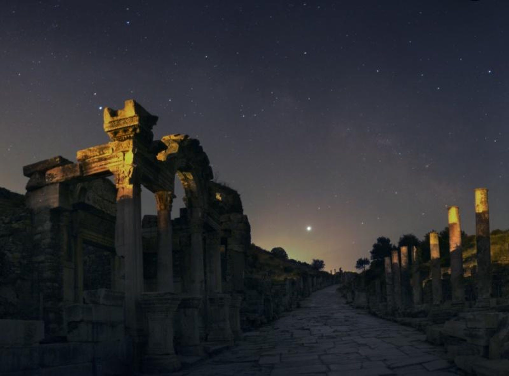 A stunning night sky panorama over the ancient ruins of Ephesus in Turkey, featuring the full moon and Jupiter at opposition, shining brightly above the southeastern horizon. The arc of the Milky Way is visible in the sky, and the ruins of the Temple of Hadrian are visible in the foreground.