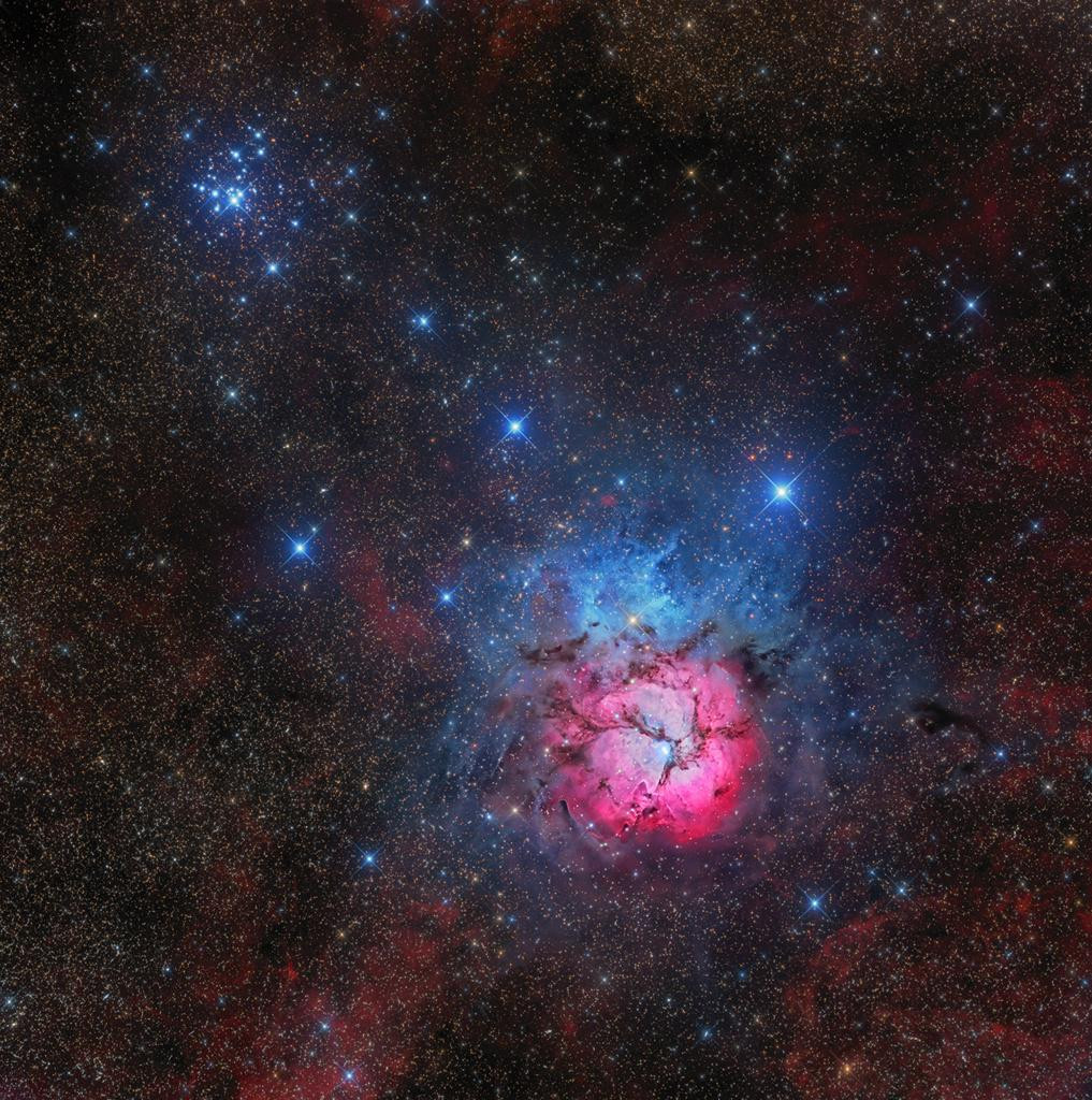 A stunning wide-field image of the Trifid Nebula (M20), a star-forming region in the constellation Sagittarius, with the open star cluster Messier 21 visible at the top left. The Trifid Nebula is a young star-forming region, with bright blue and pink emission, surrounded by dark dust lanes.