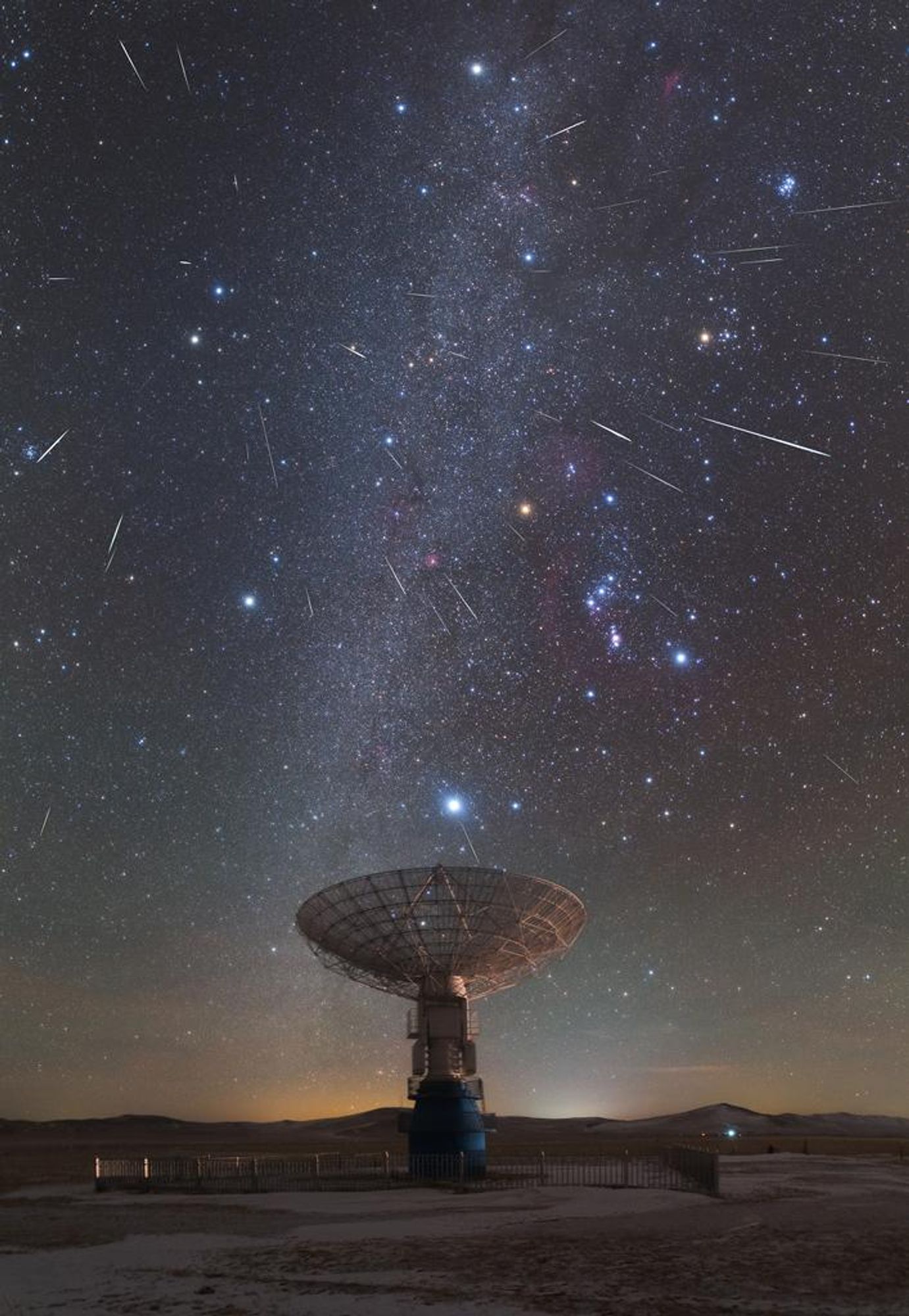 A stunning composite image of the Geminid meteor shower captured over the MUSER radio telescope in Inner Mongolia, China. The Milky Way stretches across the sky with numerous meteors streaming away from the radiant in Gemini, the constellation of the Twins. Sirius, the brightest star in the night sky, shines brightly above the radio dish. The shower's radiant is at top left, near the twin stars Castor and Pollux in Gemini.