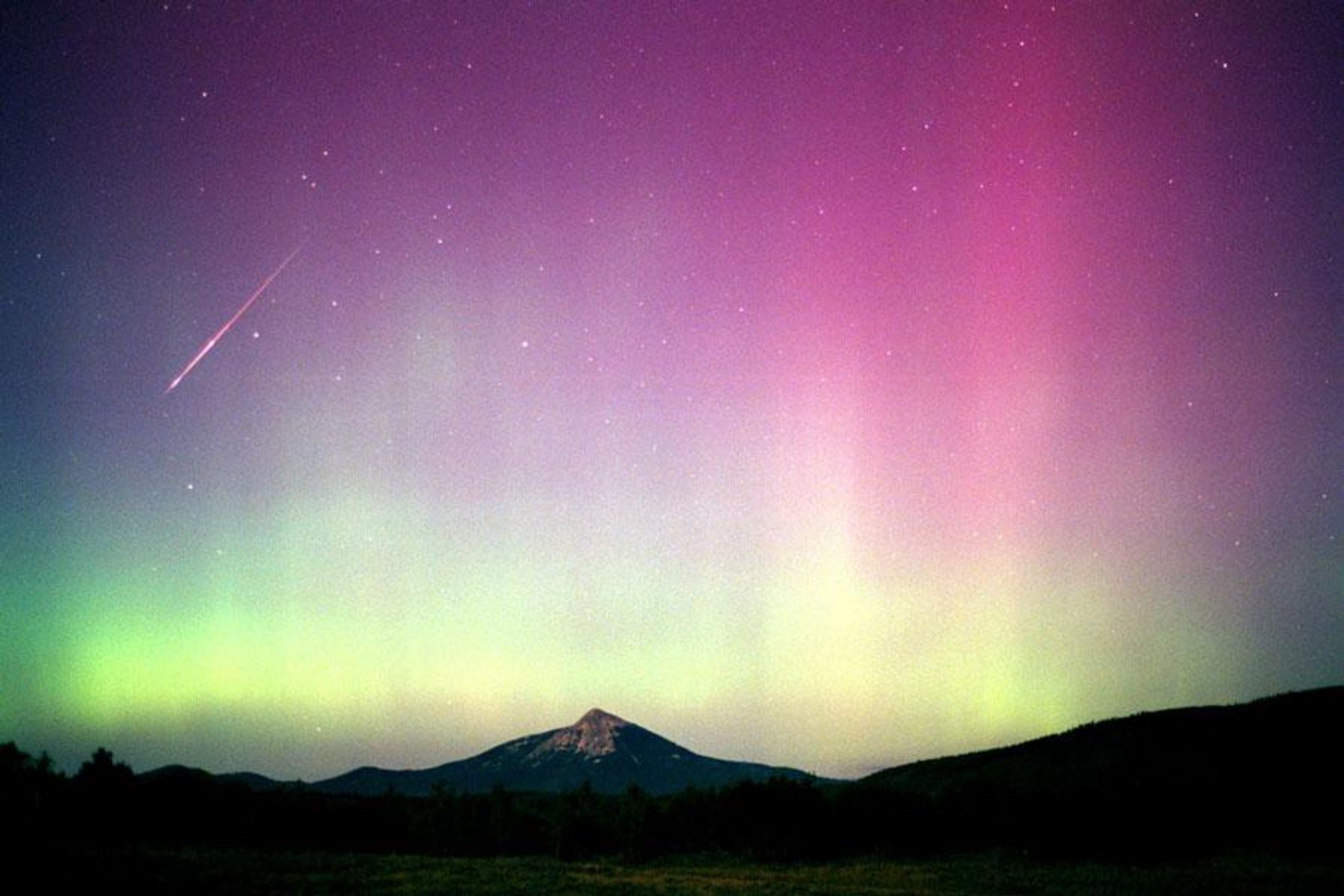 A bright Perseid meteor streaks across the sky over Colorado, captured in this photo taken in 2000. The meteor appears as a white line against a backdrop of a vibrant purple and green aurora. The landscape below is silhouetted in the foreground, with a mountain peak visible.