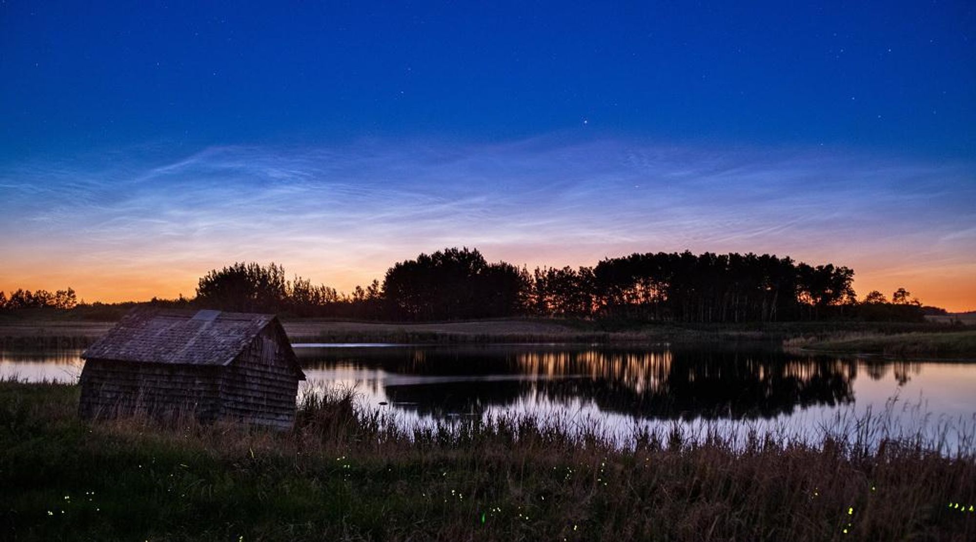 Noctilucent clouds, also known as night-shining clouds, glow in the twilight sky above a still lake in Forrest, Manitoba, Canada. The clouds appear during the summer months and reflect sunlight even after the sun has set below the horizon. Fireflies light up the foreground of this image, creating a magical scene under the summer sky.